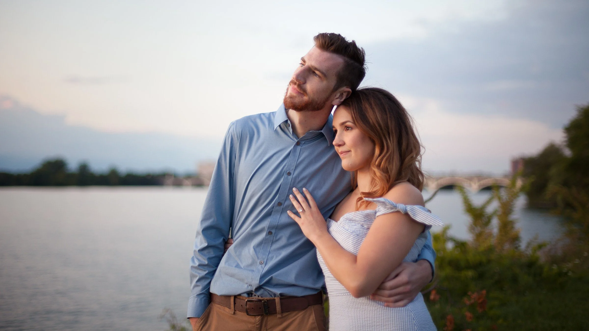 A man and woman standing close together outdoors near a body of water, with the man’s arm around the woman, both gazing into the distance at sunset.