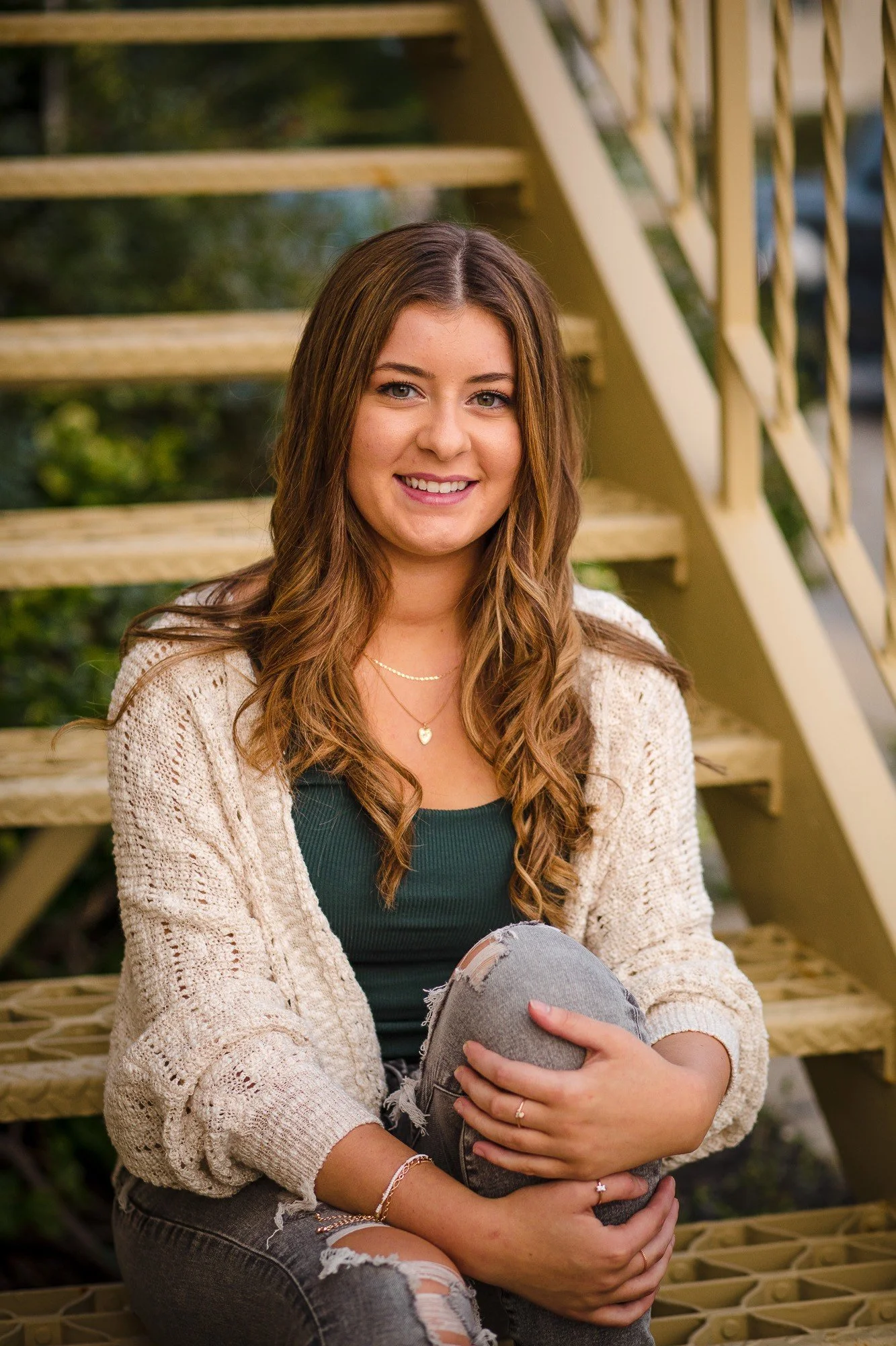 A young woman with long, wavy brown hair smiling while sitting on outdoor wooden stairs, wearing a cream cardigan, green top, and ripped jeans, with greenery in the background.