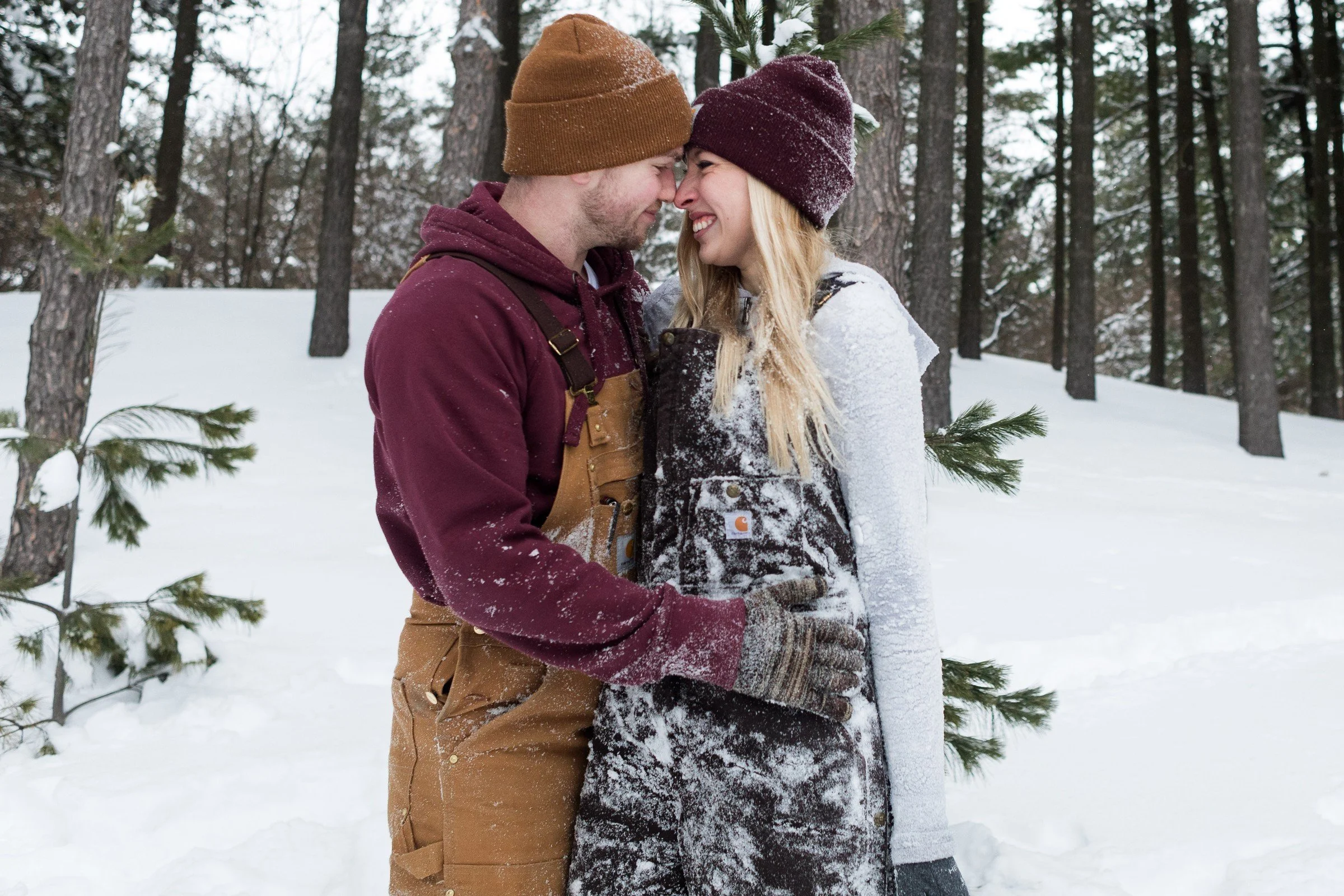 A couple smiling and holding each other in a snowy forest, wearing winter clothing and beanies.
