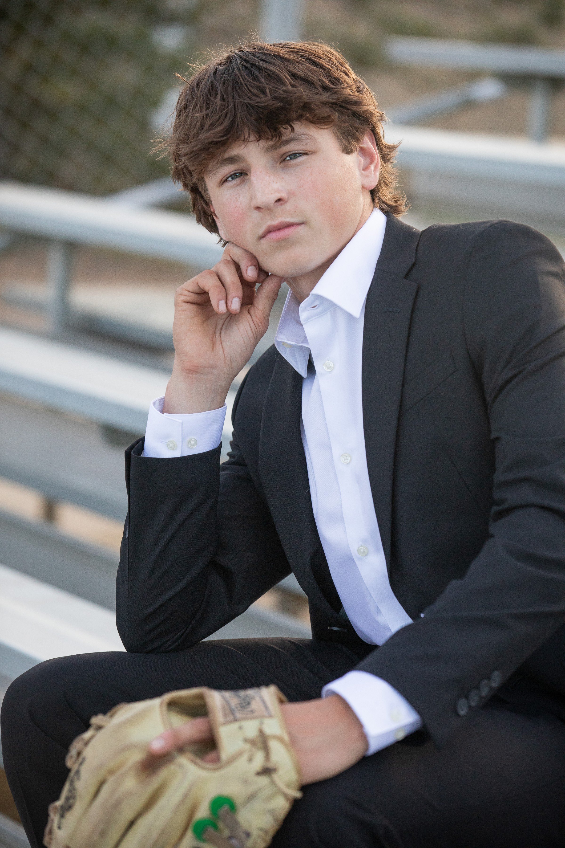 Young man in a black suit and white shirt sitting on bleachers, holding a baseball glove, with a contemplative expression.