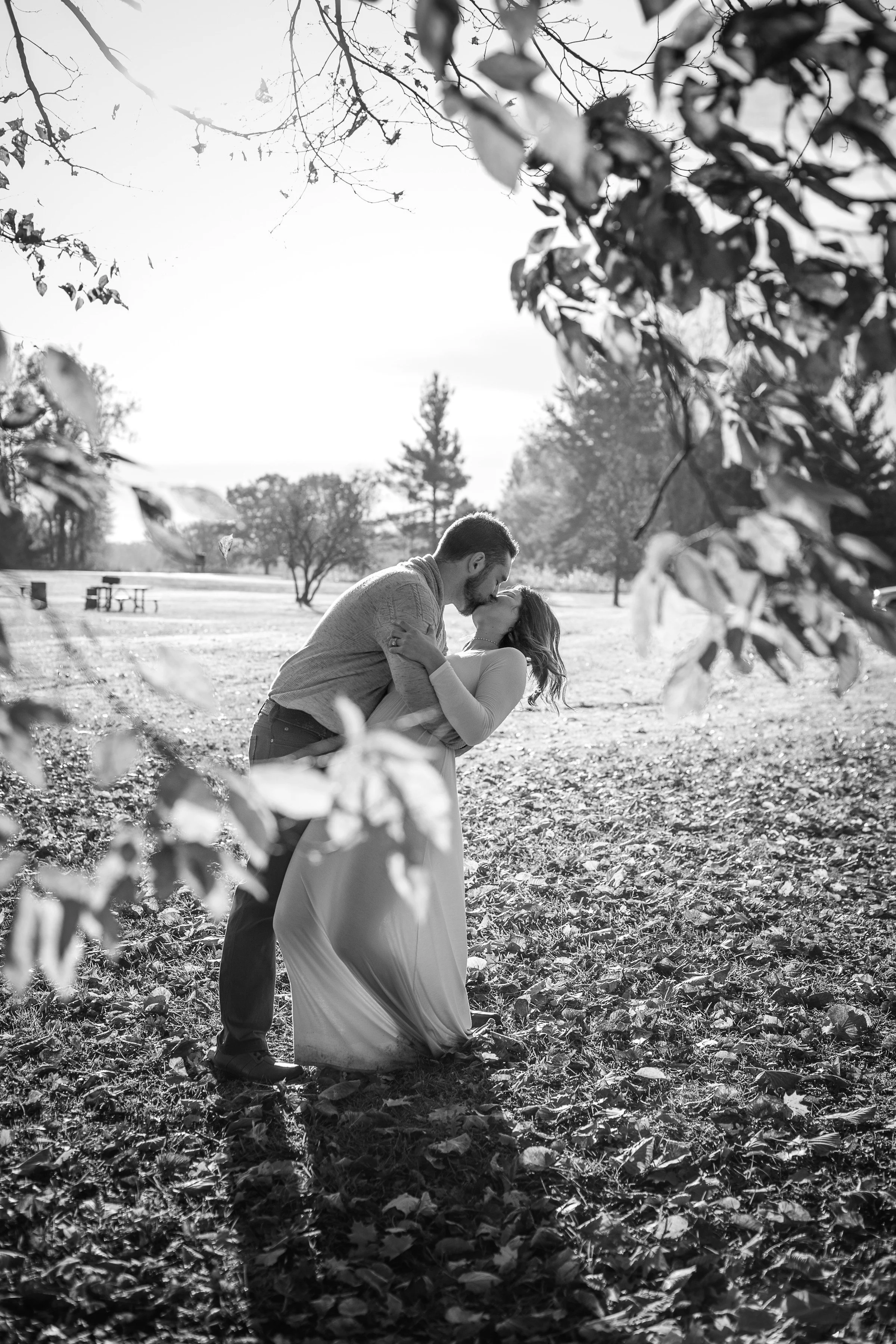 A black and white photo of a couple kissing in a park, surrounded by trees and fallen leaves, with sunlight filtering through the branches.