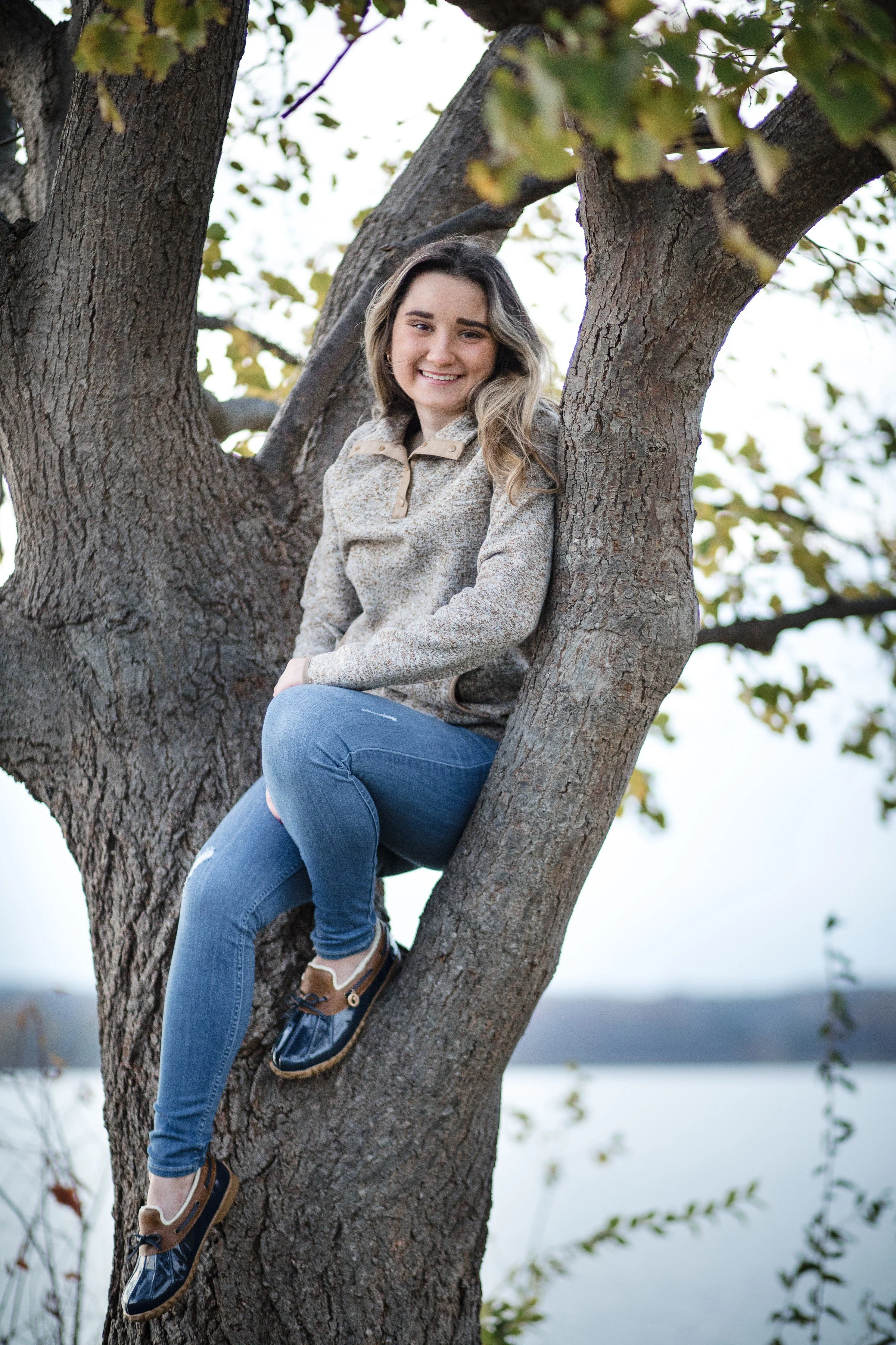 A young woman sitting in a tree near a body of water, smiling at the camera.
