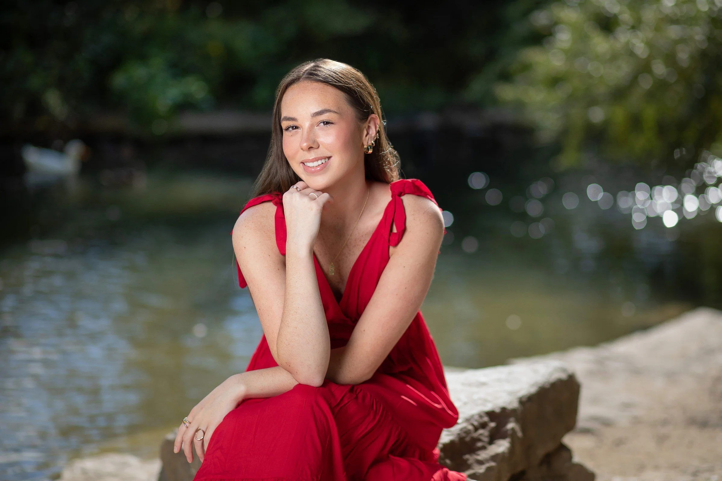 A young woman in a red dress sitting on a rock near a body of water, smiling, with a background of greenery and bokeh sunlight reflection.