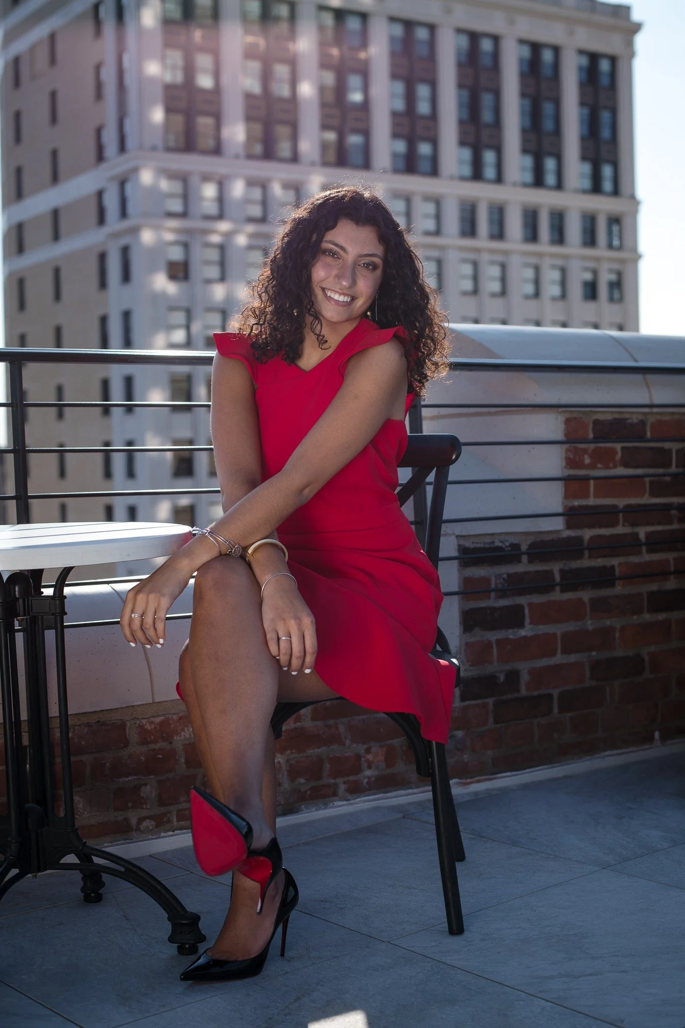 Young woman with curly hair smiling, sitting on a chair on a rooftop patio, wearing a red dress and black high heels, with a city building in the background.
