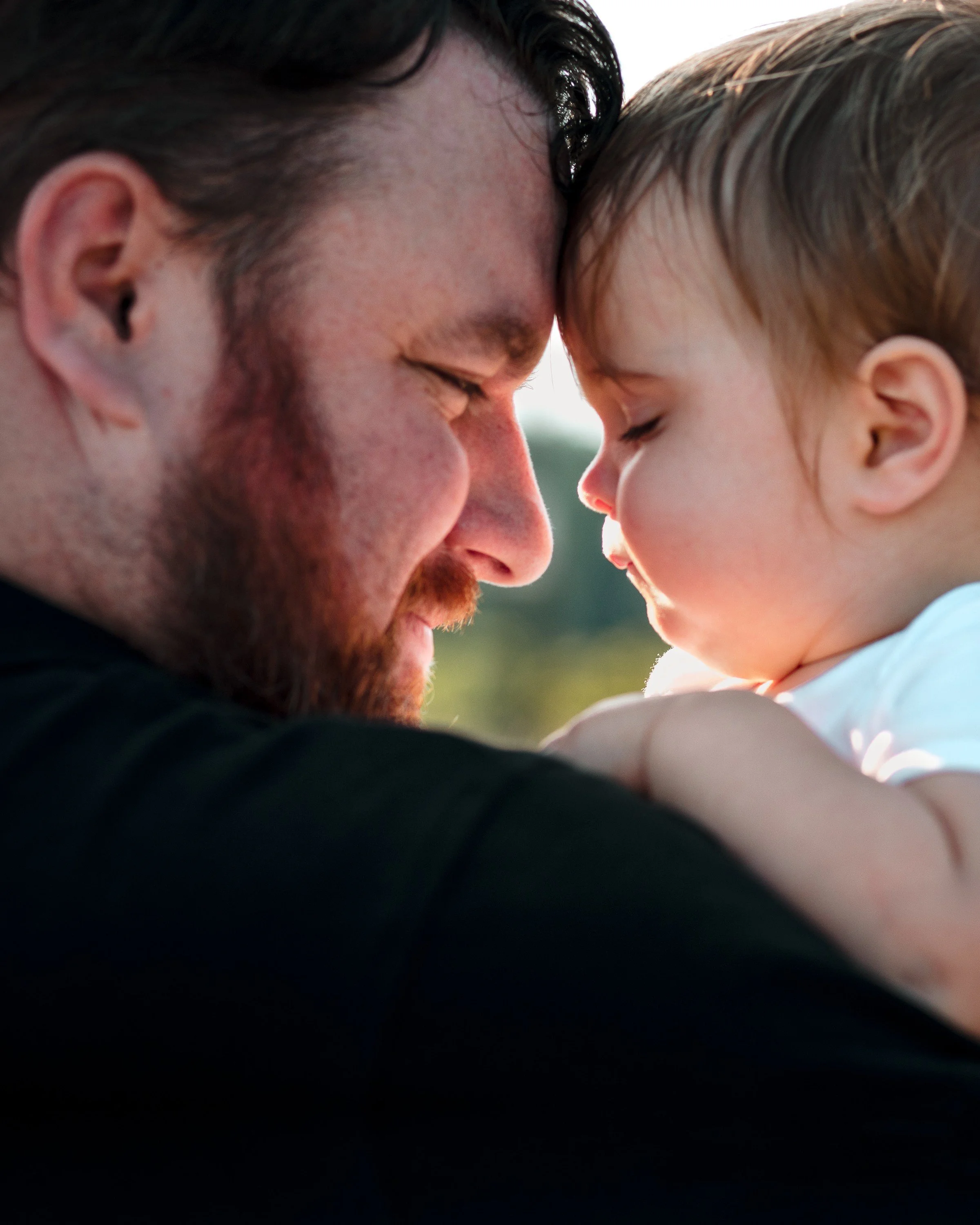 A man and a young girl with their foreheads touching, eyes closed, sharing a tender moment outdoors.