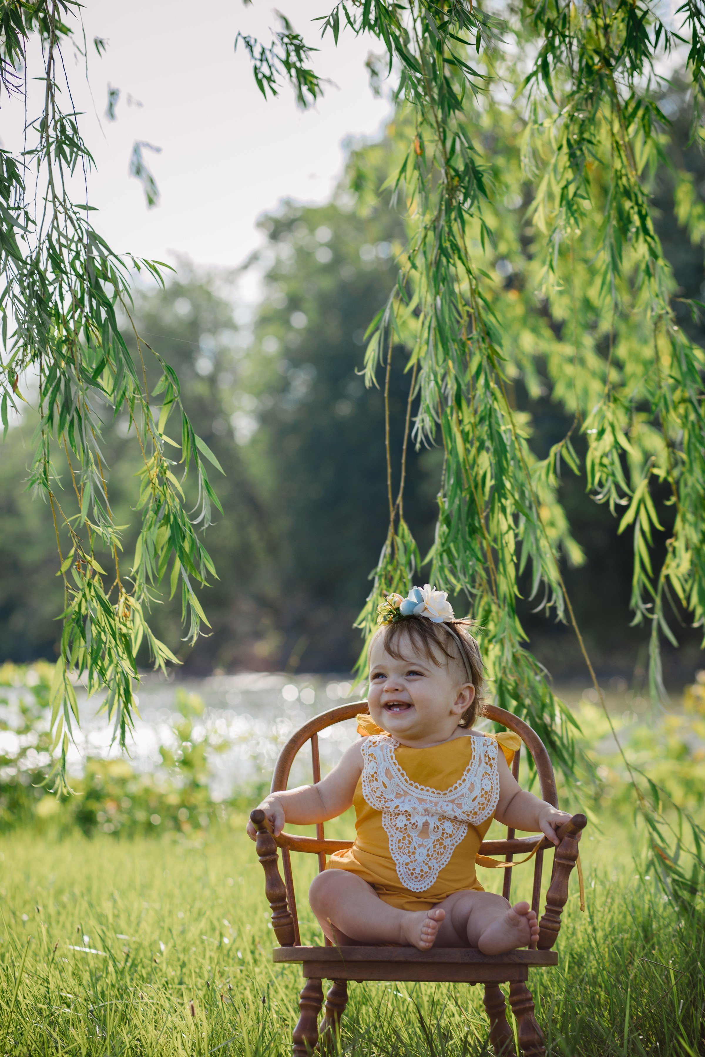 A smiling baby girl sitting on a wooden chair outdoors under hanging willow tree branches, with a river and trees in the background on a sunny day.