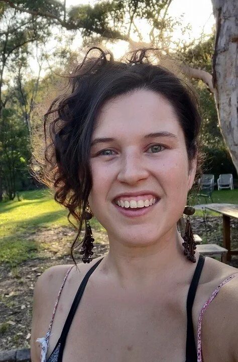 A woman with short, curly dark hair smiling outdoors in a park with trees and a bench in the background.