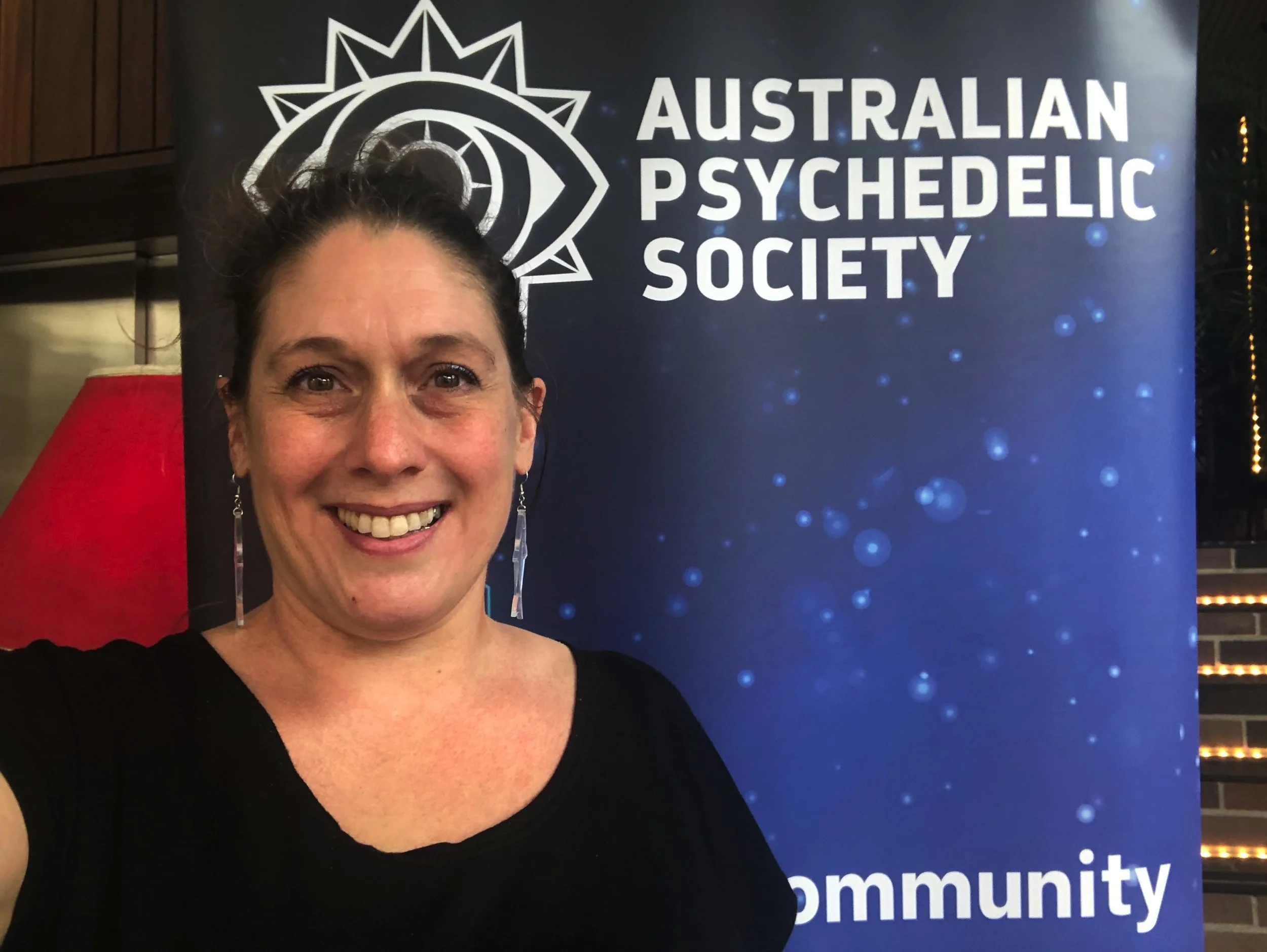 A woman with dark hair and earrings smiling in front of a blue banner that reads 'Australian Psychedelic Society'. The banner features a logo with a stylized eye and sunburst design.