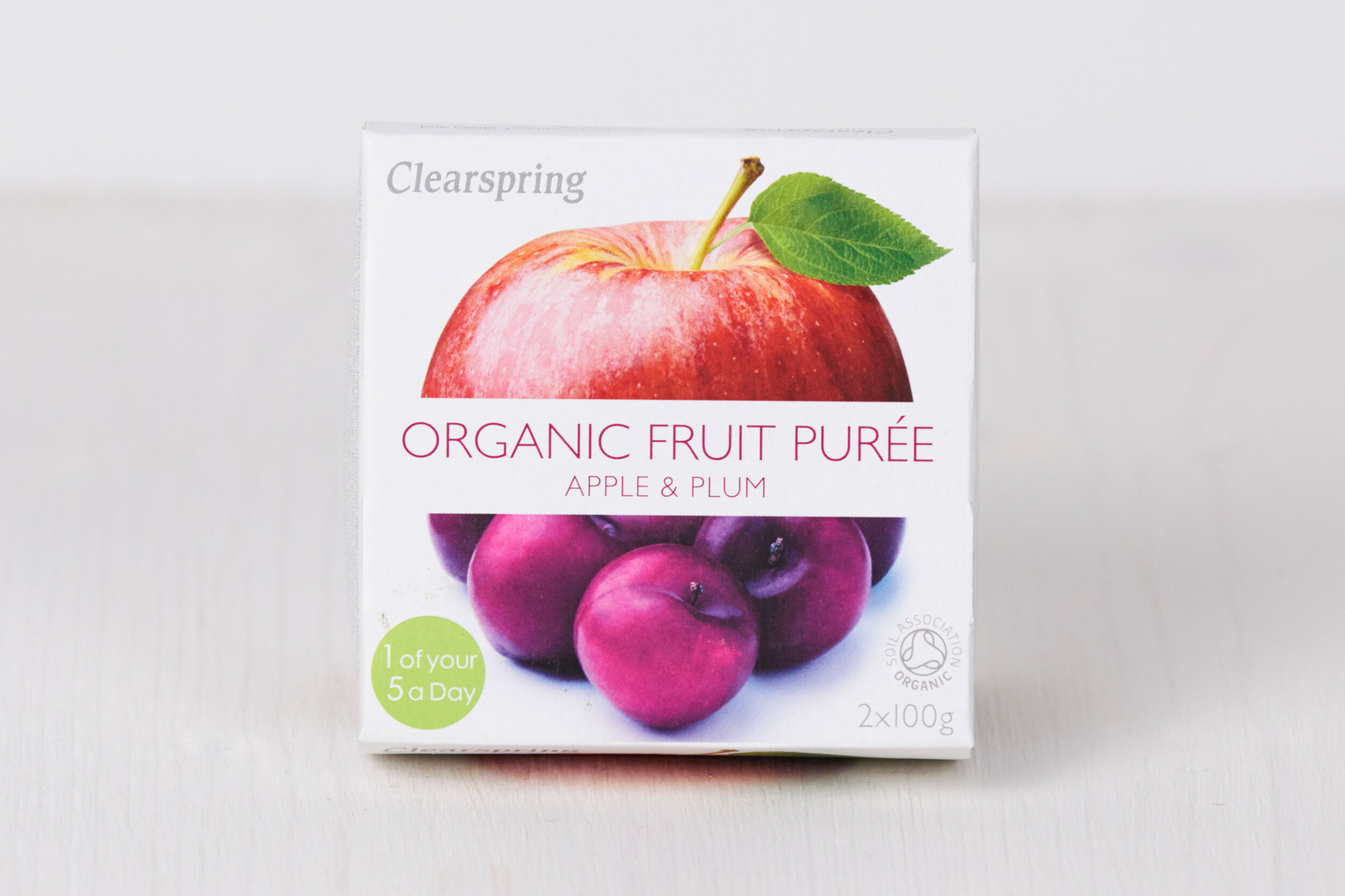 Box of Clearspring organic fruit purée with apple and plum, featuring an image of an apple with a leaf and a bunch of plums on the front.