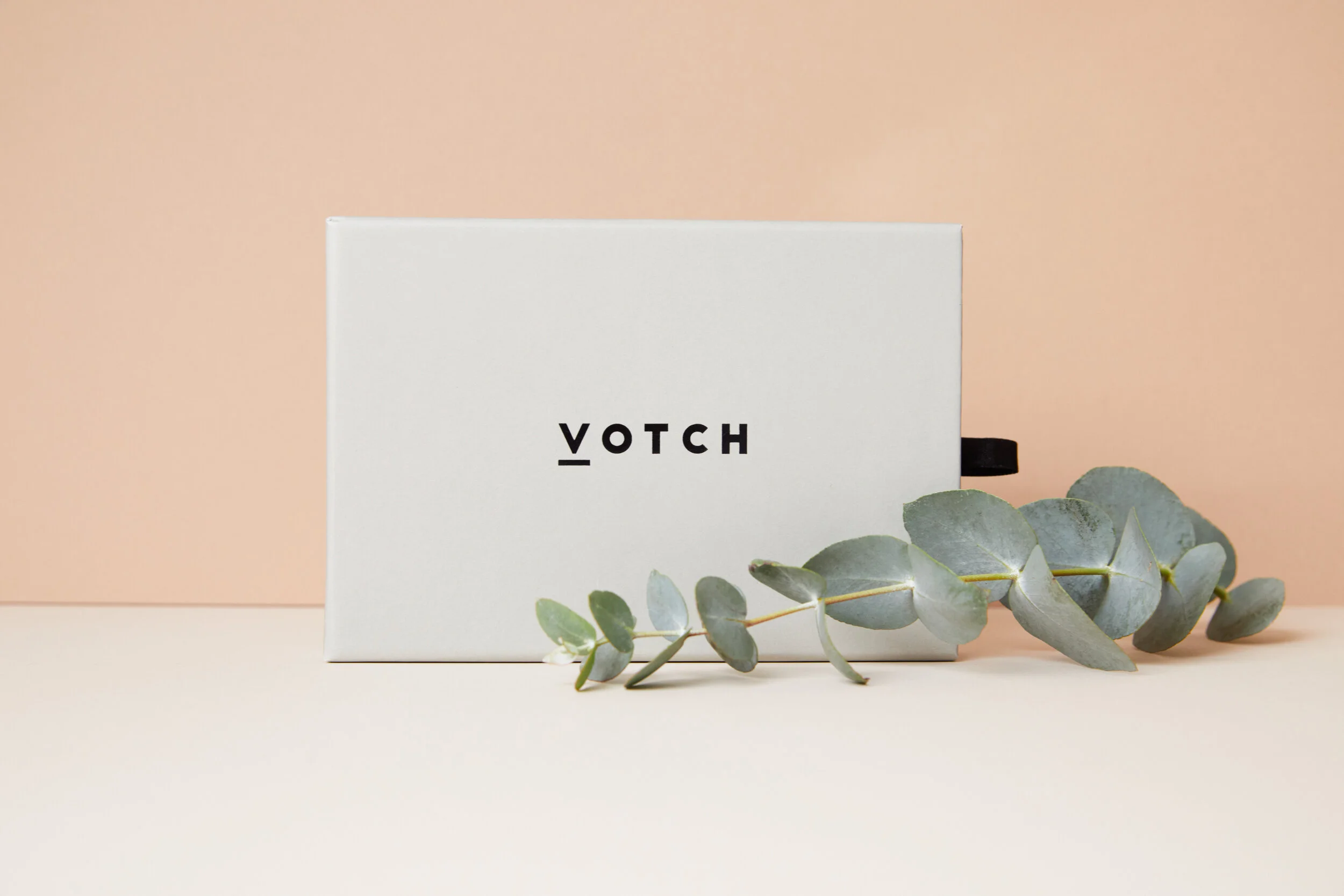 A white box with black text 'VOTCH' on it, placed on a beige surface with a eucalyptus branch in front, against a pink background.