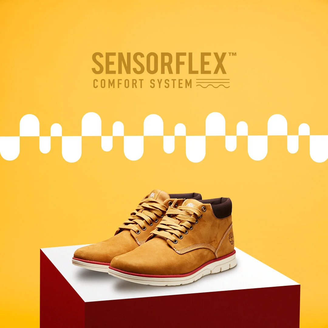 A pair of tan leather Timberland boots displayed on a red and white platform against a yellow background with the text 'SensorFLEX Comfort System' above.
