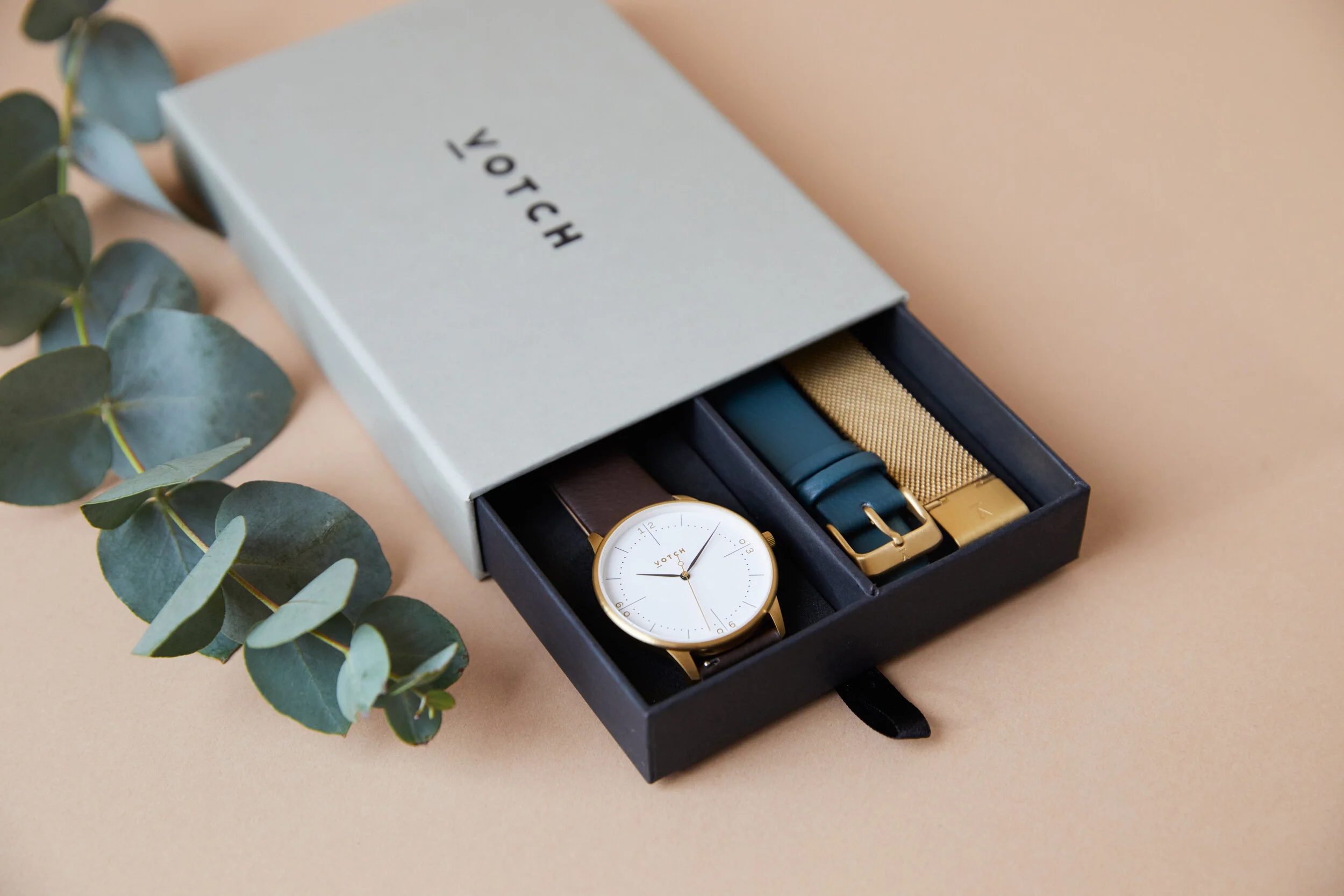 A watch box with four watch bands and a watch, moving from left to right, with a eucalyptus branch to the left on a beige background.