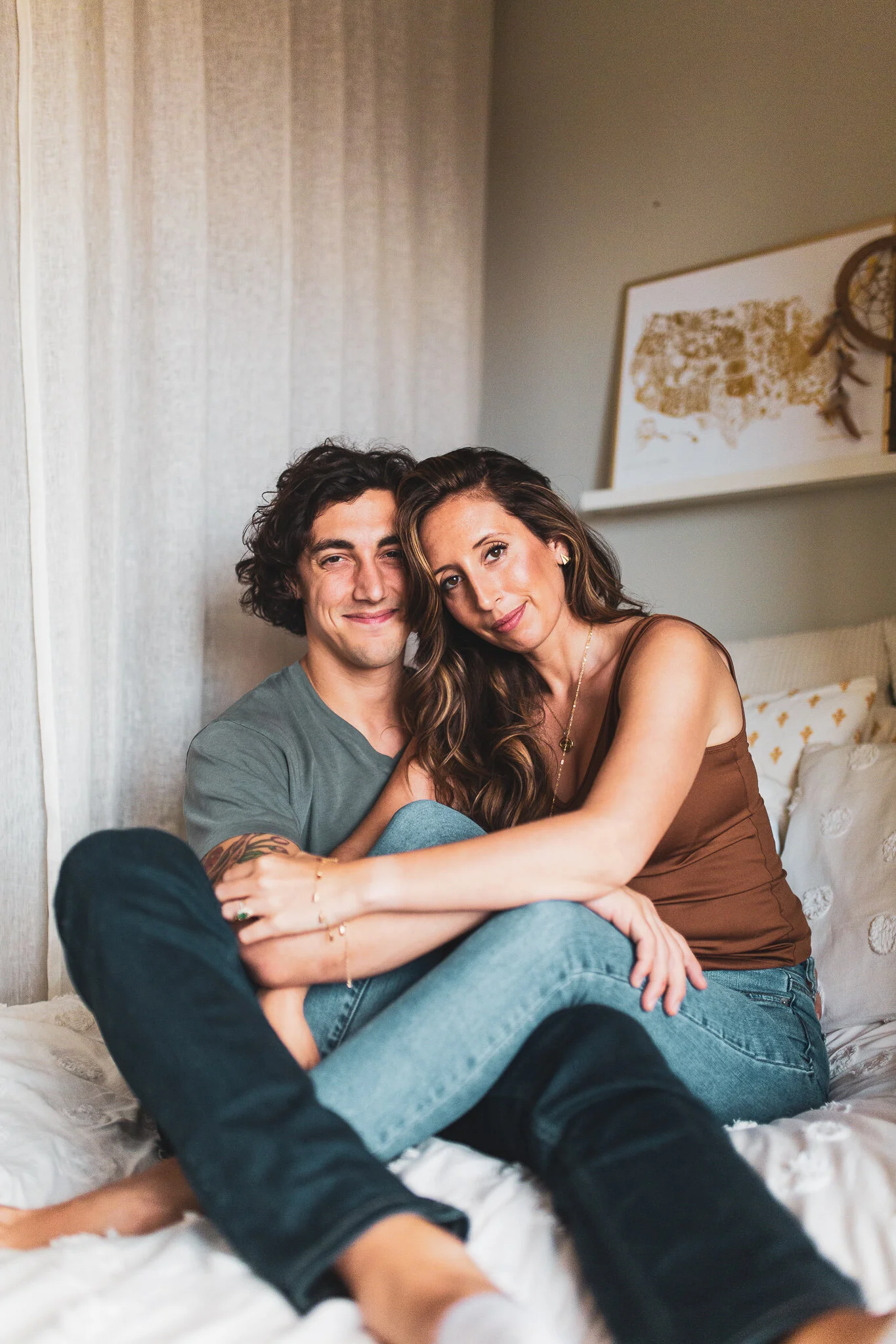 A couple sitting on a bed, embracing and smiling, in a cozy room with minimal decor including a framed map and dreamcatcher on a shelf.