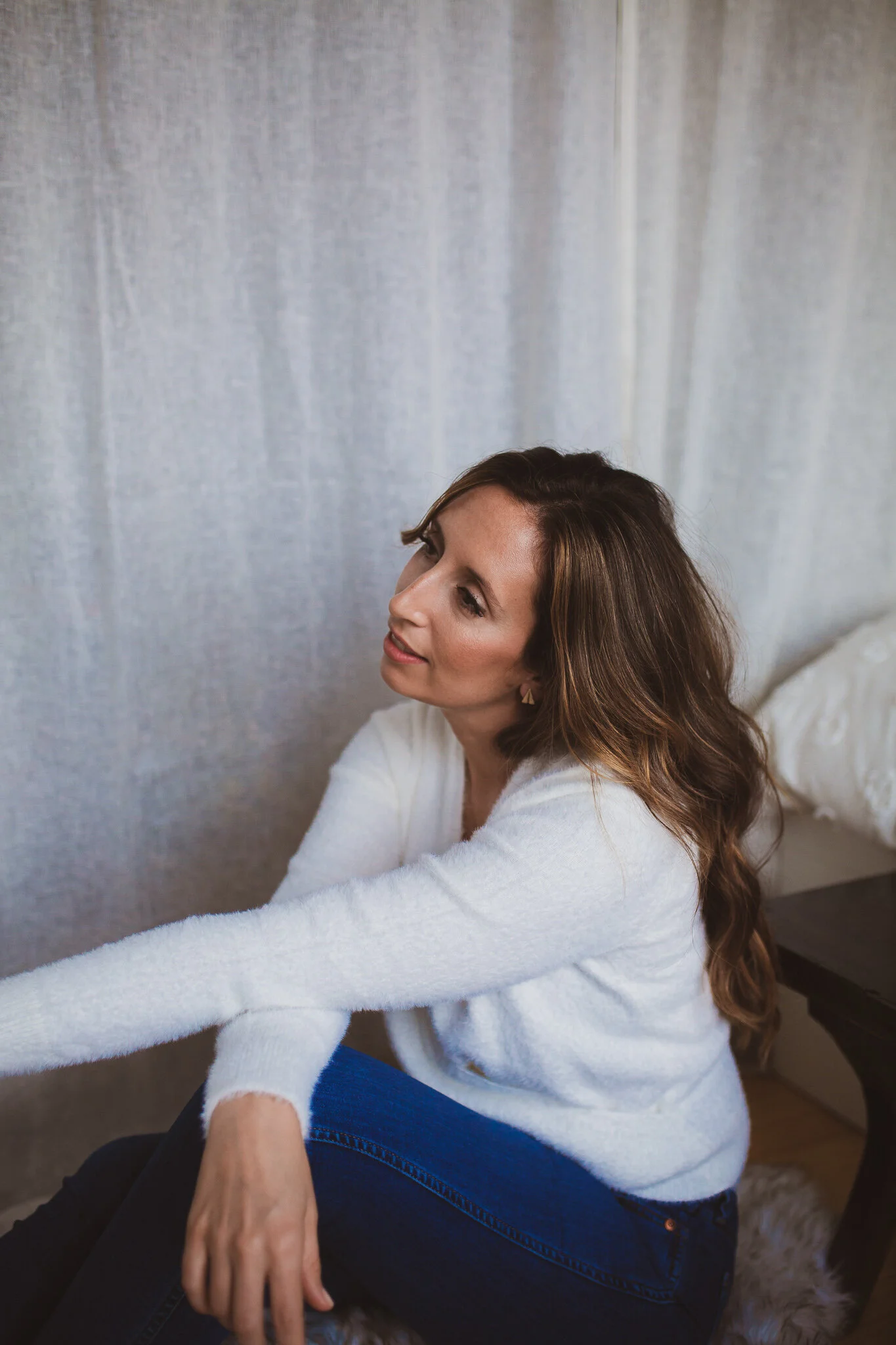 A woman with long brown hair wearing a white sweater and blue jeans sitting on a furry stool against a gray curtain background.