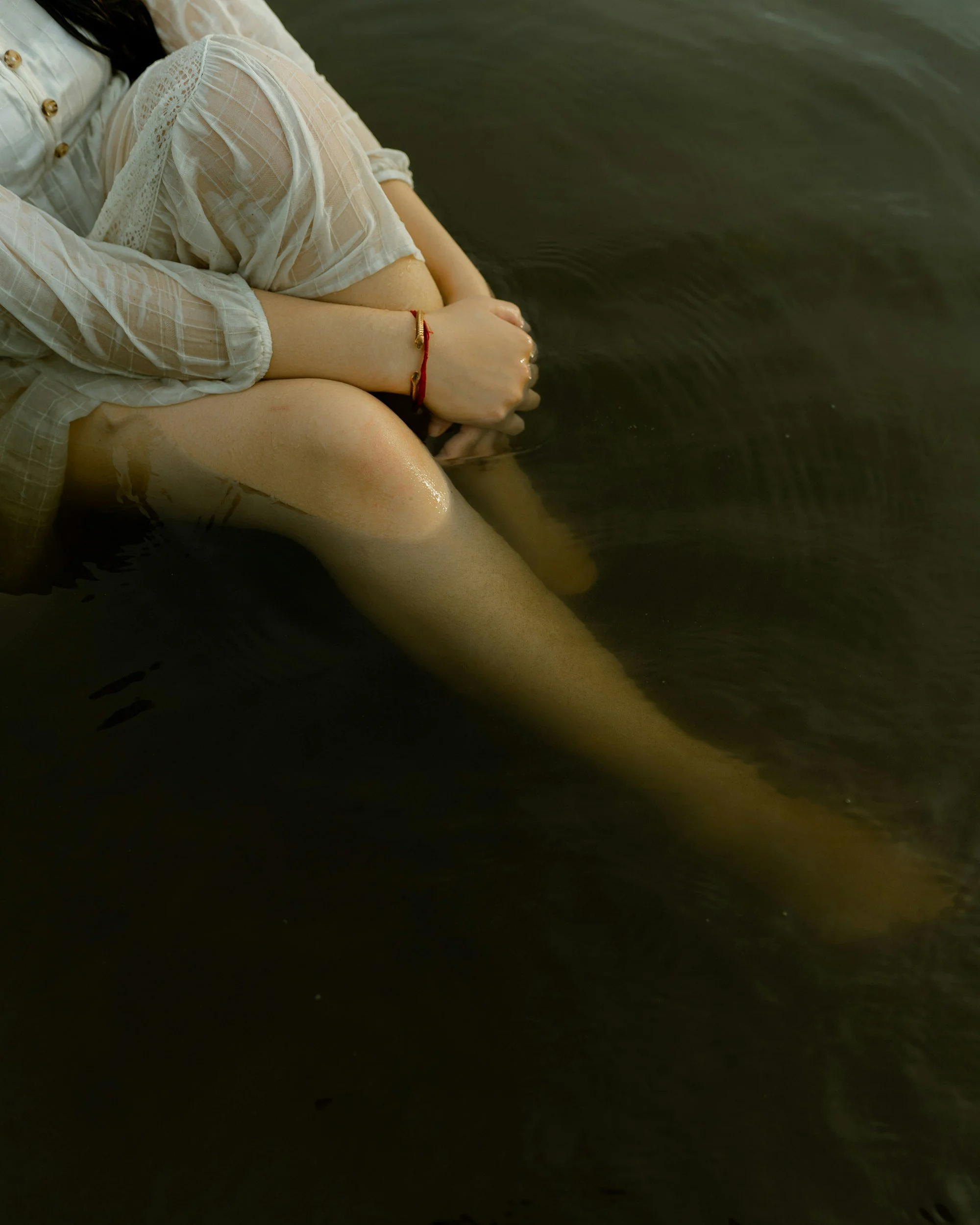 A person sitting in water, their knees drawn up, wearing a light-colored, sheer, long-sleeved blouse and a red bracelet on their wrist.