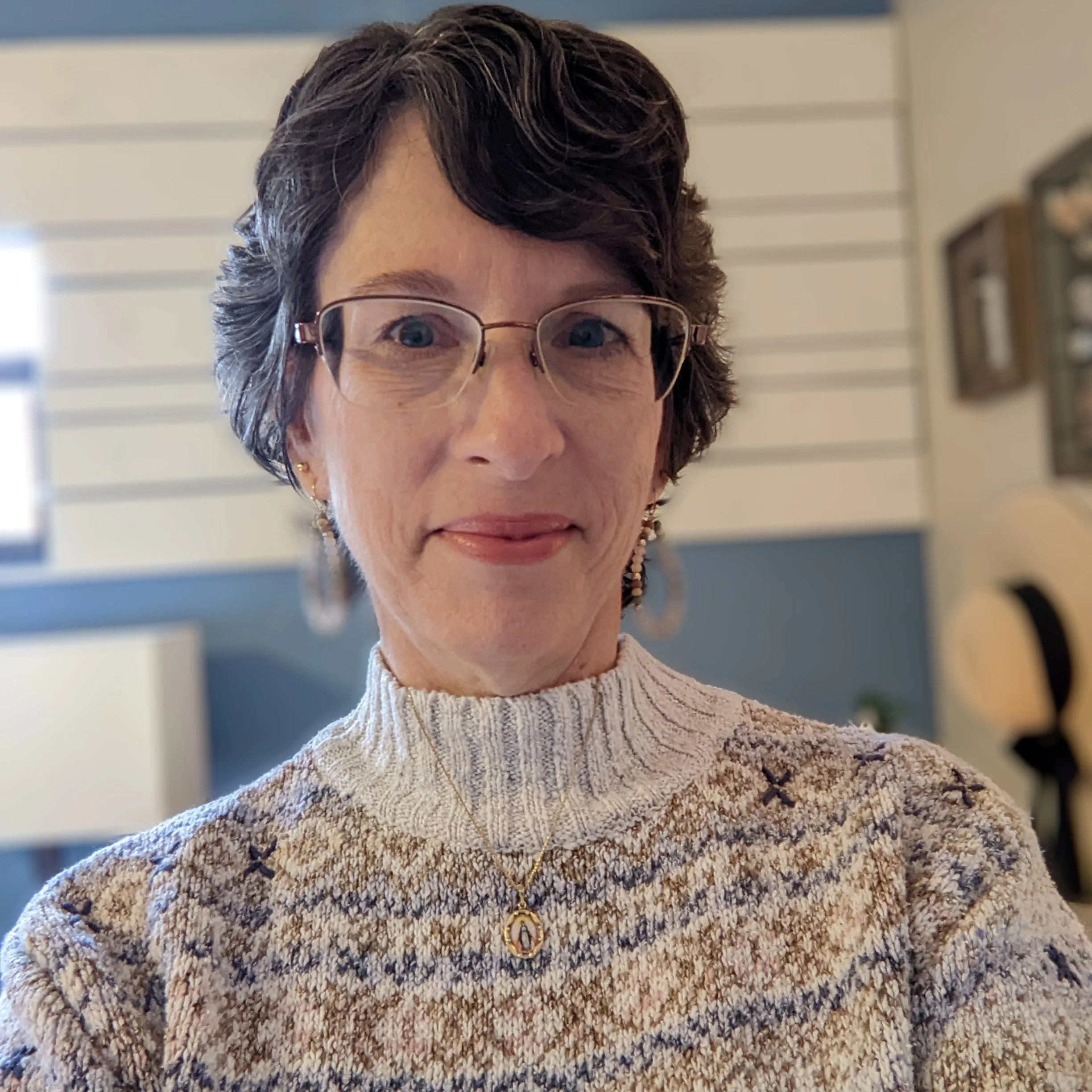 A woman with short dark hair, wearing glasses, earrings, a beige patterned sweater, and a gold necklace with a religious pendant, smiling indoors.