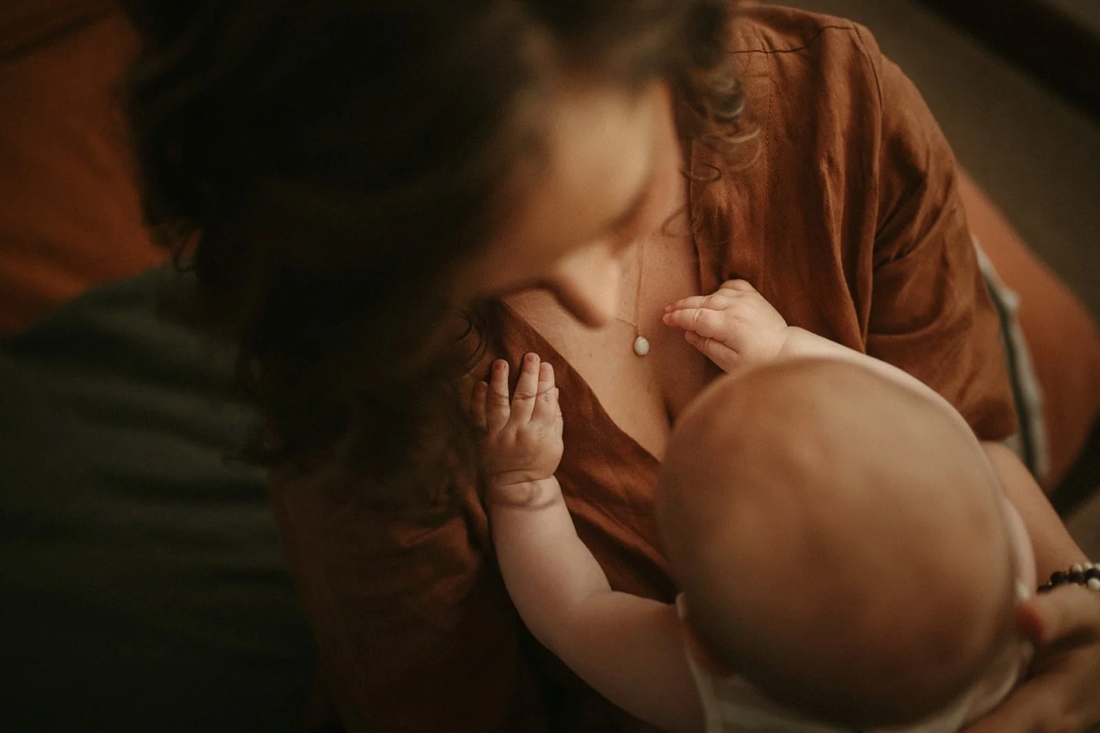 A woman with long curly hair, wearing a brown top, is breastfeeding a baby who is lying on her chest. The baby's hand rests on her chest, and the woman is wearing a necklace with a white pendant.