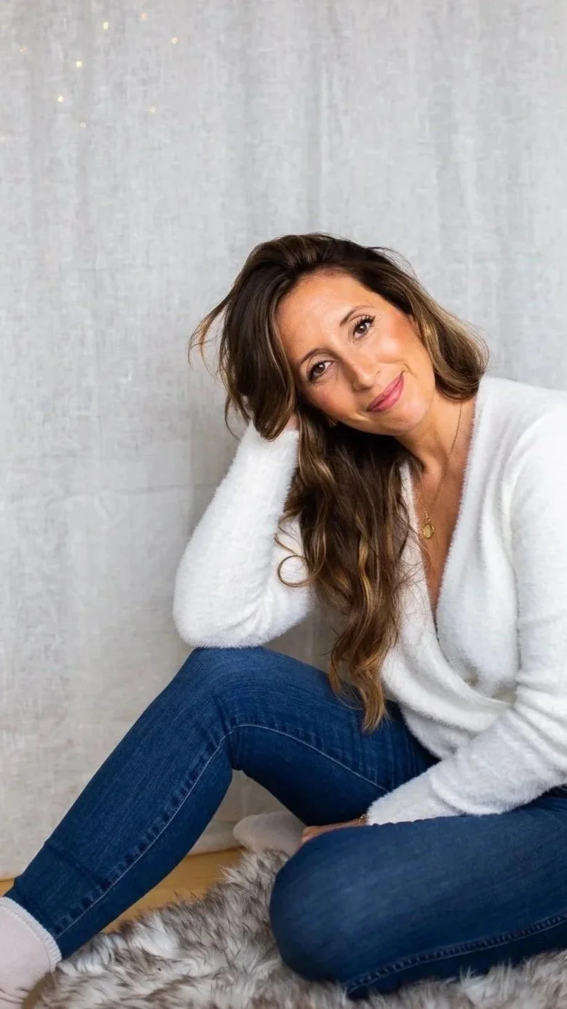 A woman sitting on a fluffy rug against a light-colored wall, smiling and resting her head on her hand, with long wavy brown hair, wearing a white sweater and blue jeans.