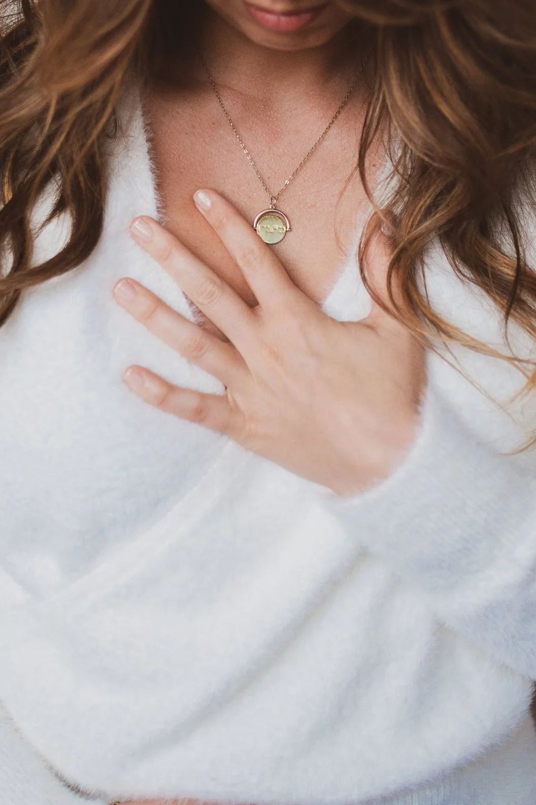 Close-up of a woman wearing a white sweater, with her hand on her chest, showing a necklace with a rainbow pendant with a small tree design inside.