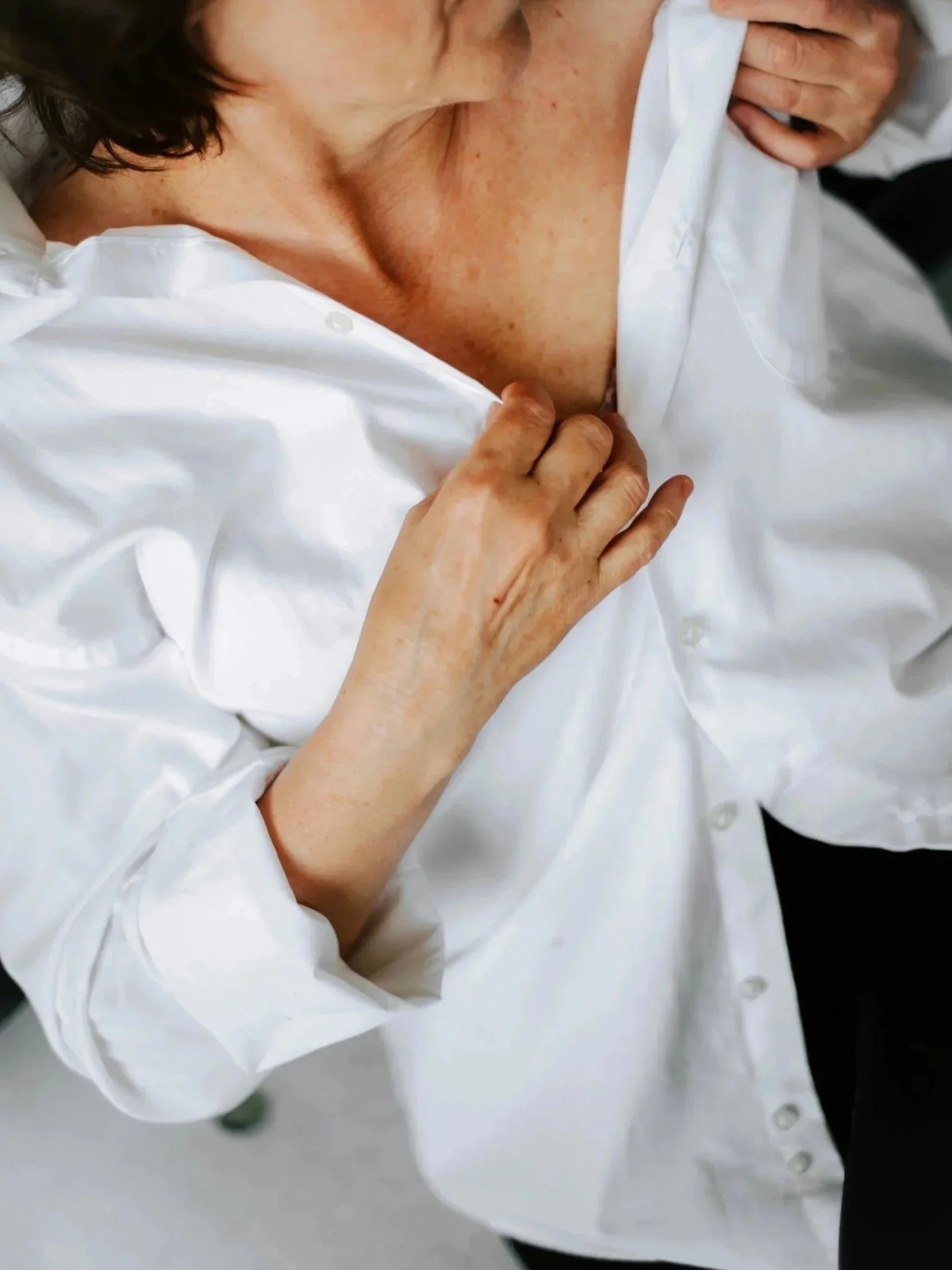 A woman with slightly tan skin and freckles is unbuttoning a white dress shirt and revealing her chest.