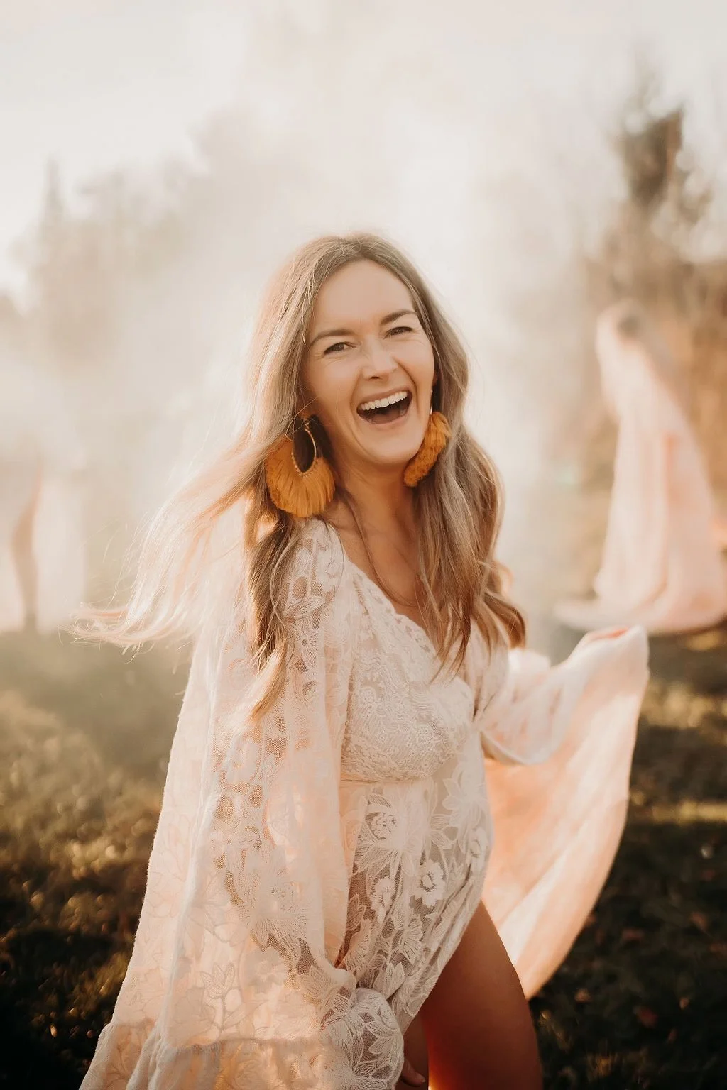 A woman with long, wavy hair wearing large orange feather earrings and a lace, flowy dress, smiling and laughing outdoors during golden hour.