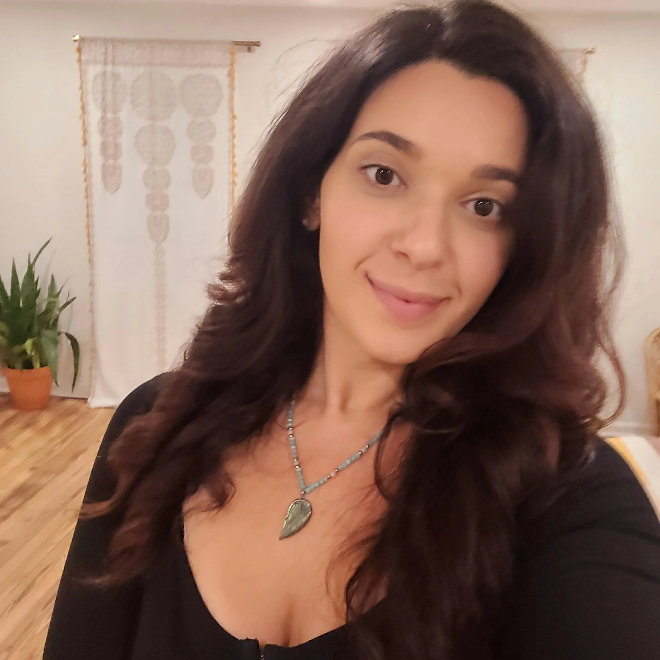 A smiling woman with long, wavy dark hair taking a selfie in a room with wooden flooring, white walls, a potted plant, and decorative curtains.