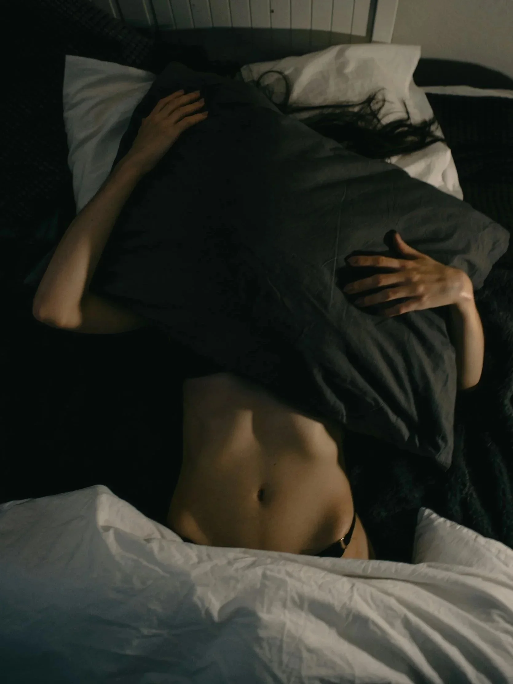 Person lying on a bed with arms crossed over their chest, wearing a black shirt and having visible midriff, with dark hair spread out on a white pillow.