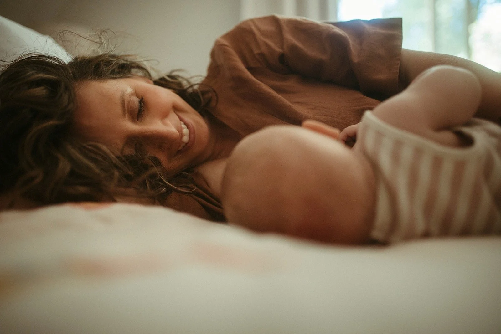 A woman with curly hair smiling and lying on her side, looking at a baby who is lying on a bed, with a window in the background.