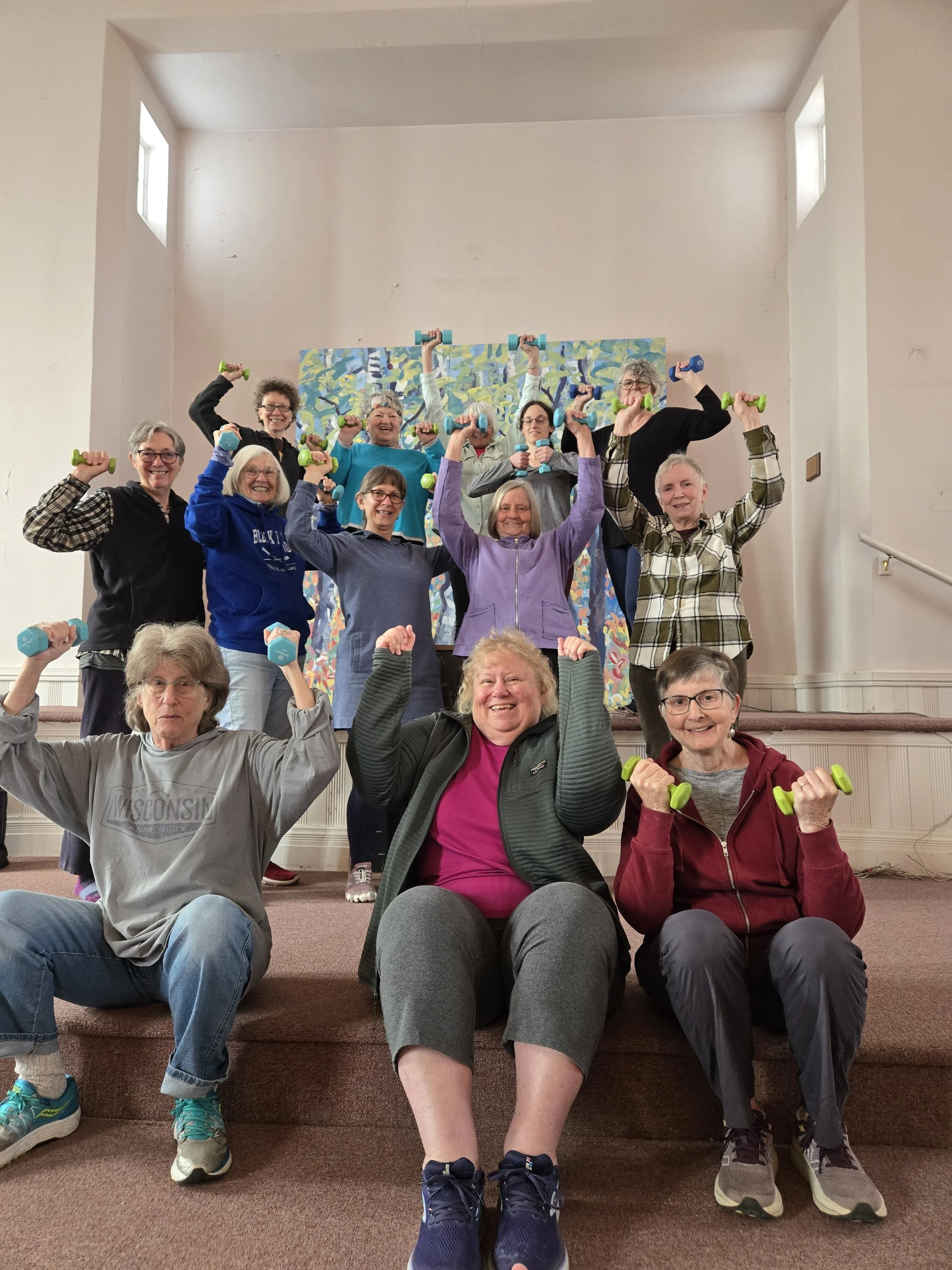 Group of elderly women holding small green and blue dumbbells, posing happily on a staircase in a room with white walls and a colorful painting behind them. Some women are sitting in front, while others are standing on stairs.
