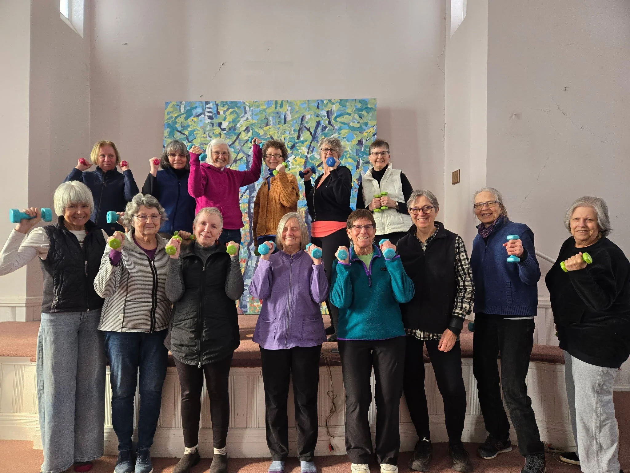 The Bone Builders Group exercising with small dumbbells inside the Westford Commo Hall. They are smiling and standing in front of a colorful abstract painting created by the community at a community gathering in our space.