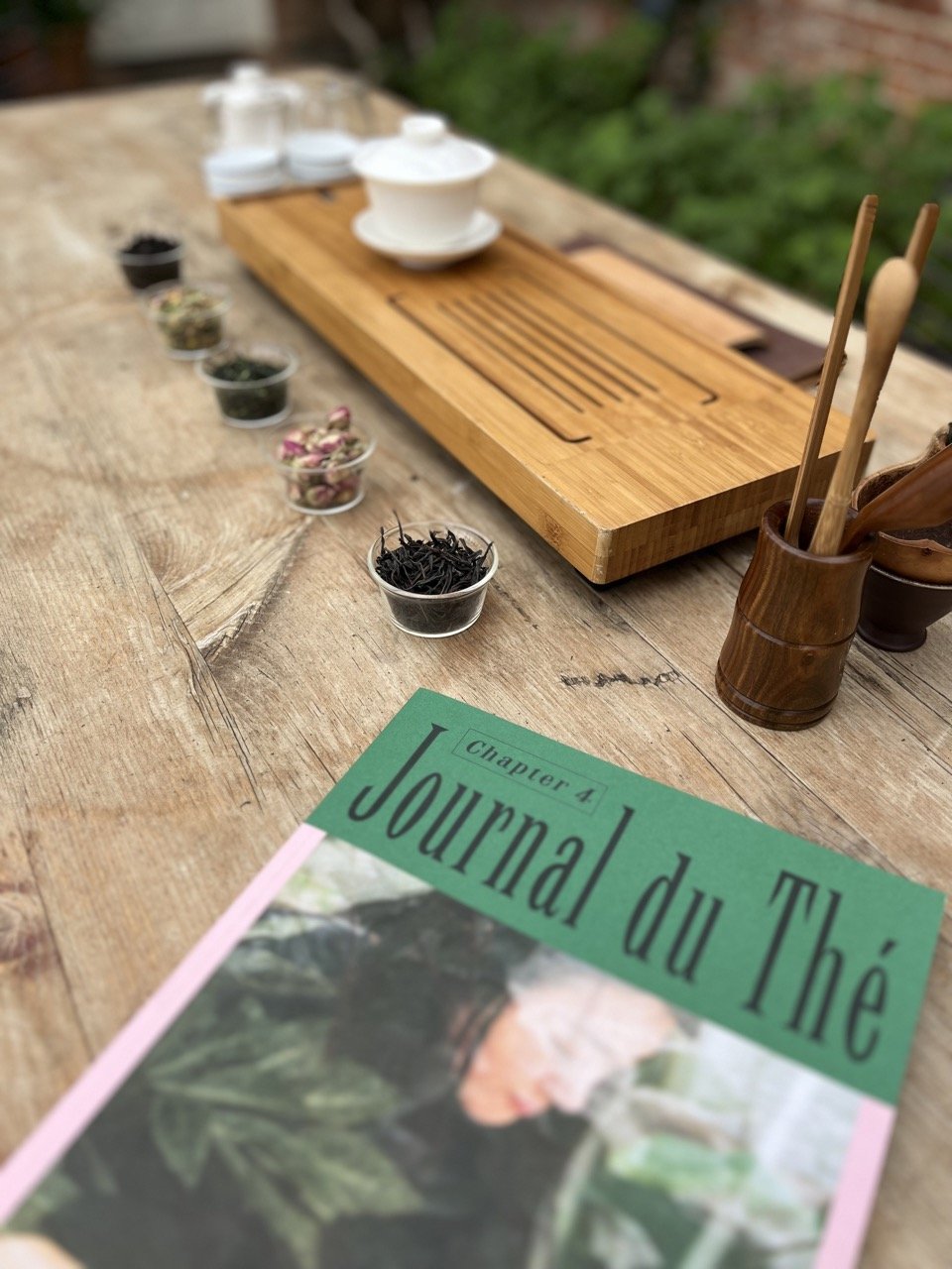 A rustic outdoor tea setup with a wooden table, a tea tray, small containers of herbs and flowers, a small cup of tea utensils, and a magazine titled 'Journald du Thé' on the wooden surface.