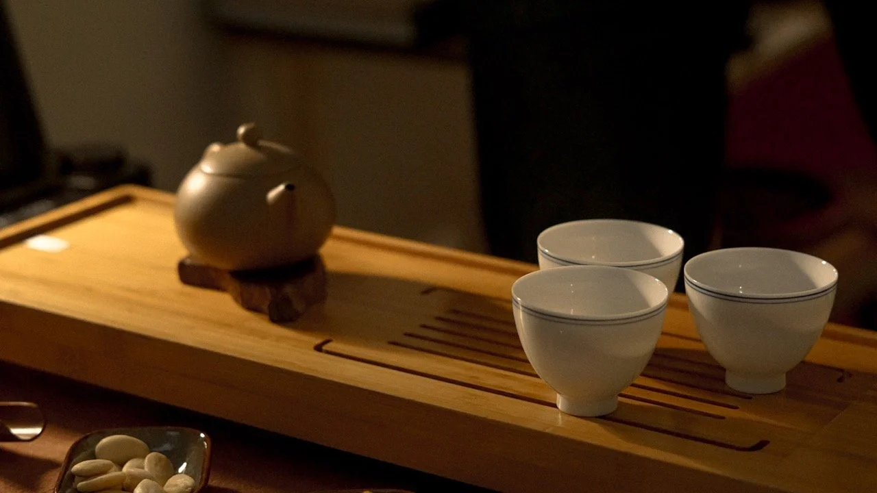 A ceramic teapot and three small white teacups on a wooden tray, with a dish of mixed nuts in the foreground.