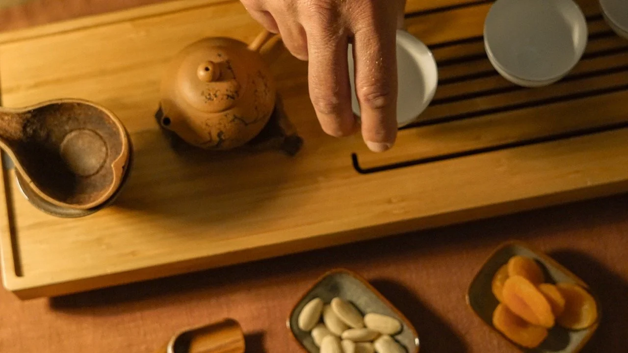 A person’s hand is pointing at a wooden tray with tea pots, cups, and bowls of snacks, including pistachios and dried apricots.