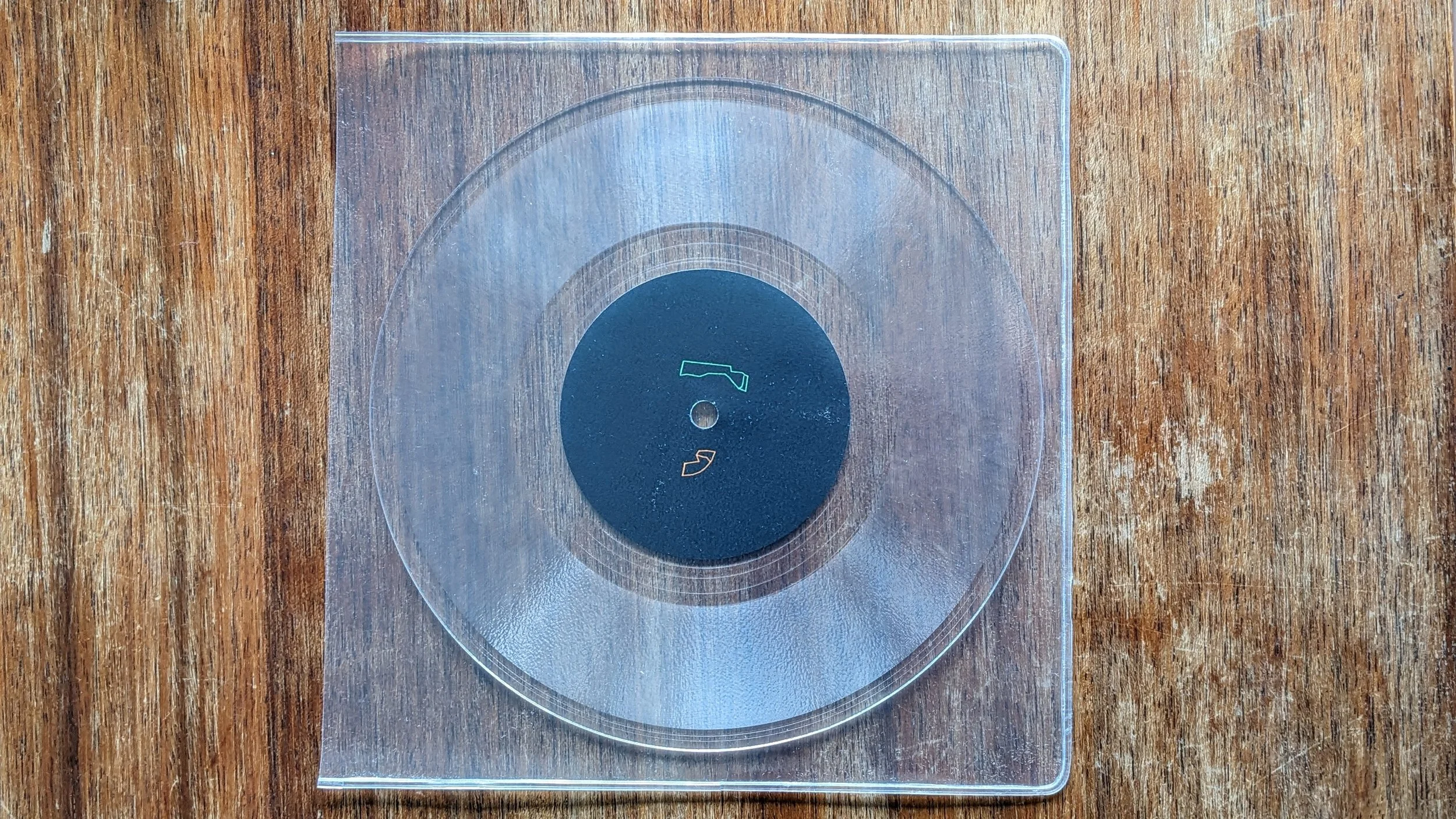 Kar Pouzi clear vinyl record on scratched table. No text on black label at centre. Instead, 2 small hooves appear, one in green, one in orange.