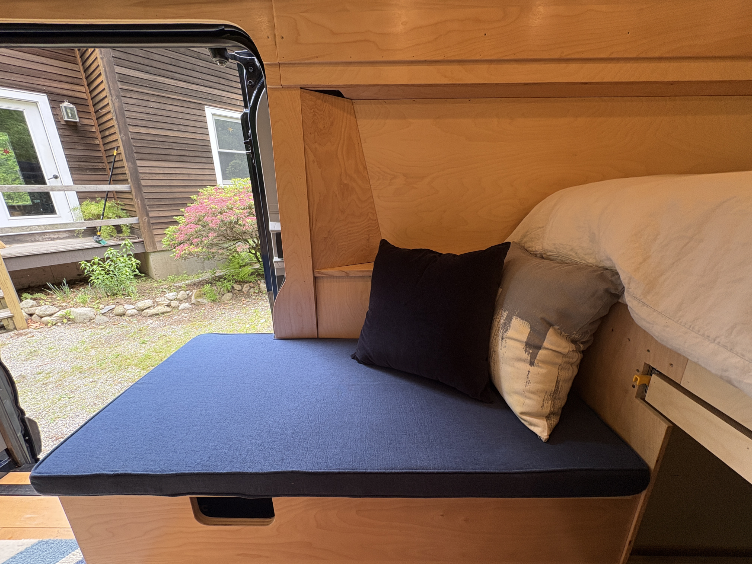 Interior of a camper van with a bed, black pillows, and an open door showing an outdoor scene with a flowering shrub, a wooden deck, and a brown house with white window frames.