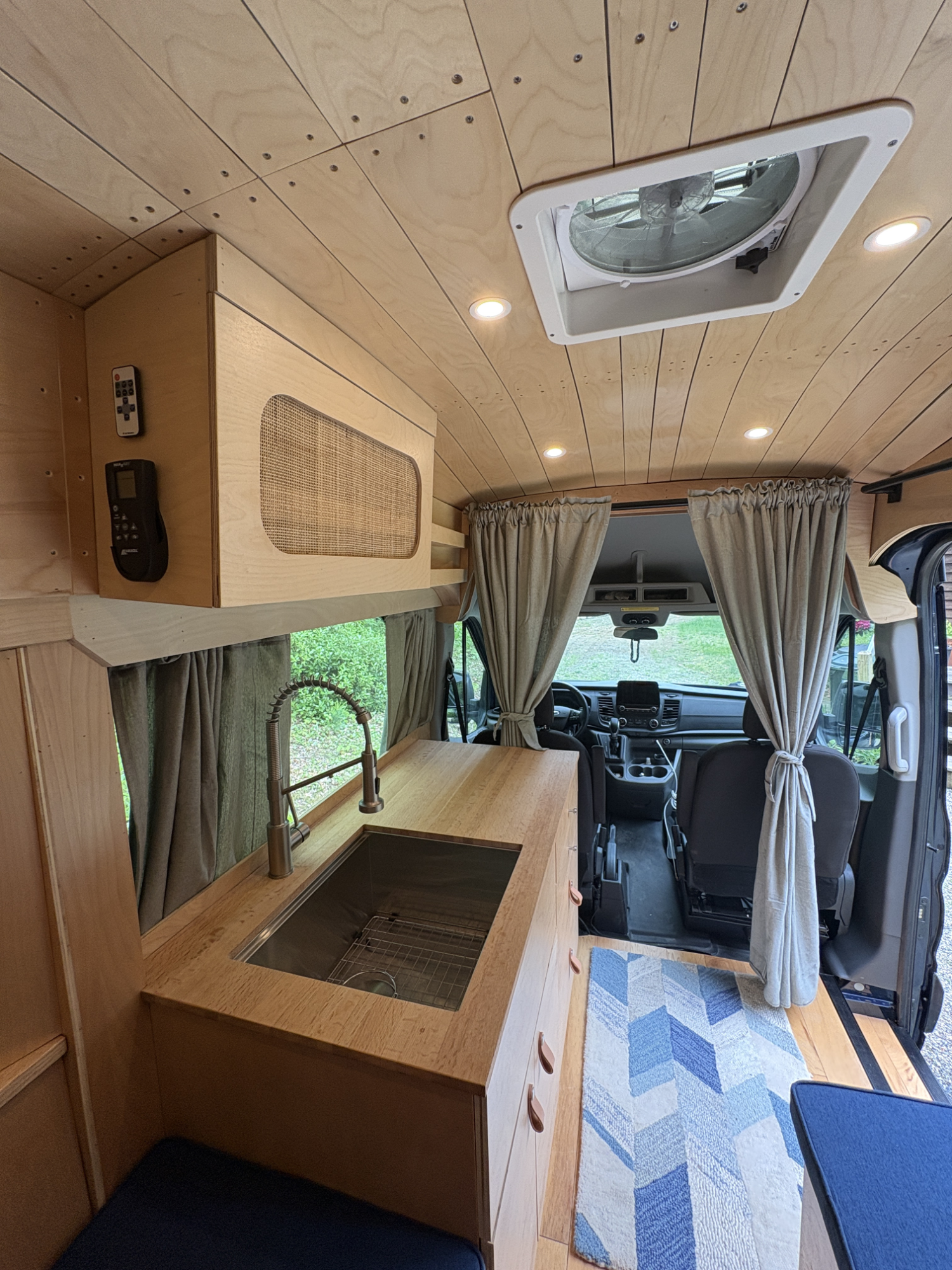 Interior of a camper van showing a kitchen area with a wooden countertop, a sink, and a faucet. There are curtains in front of the driver’s area and a blue geometric-patterned rug on the floor. The ceiling has recessed lighting and an air vent. The d