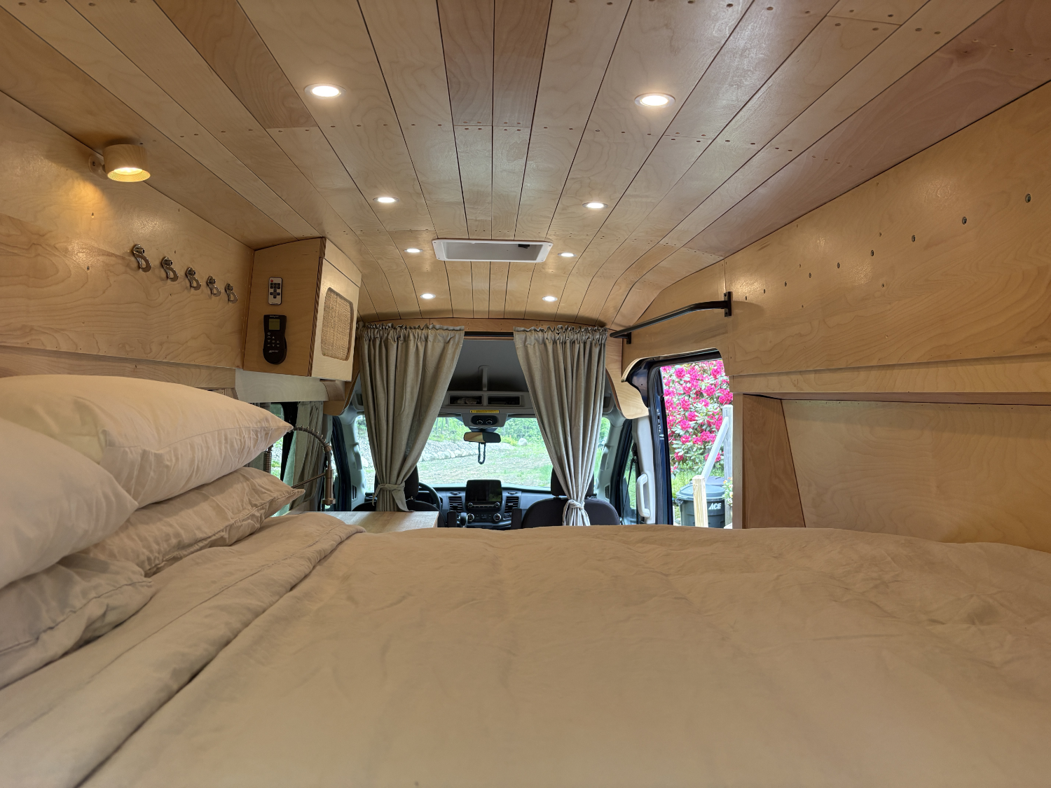 Interior of a camper van with a bed in the foreground, a window with curtains, and the dashboard and front seats visible at the back with pink flowers outside.