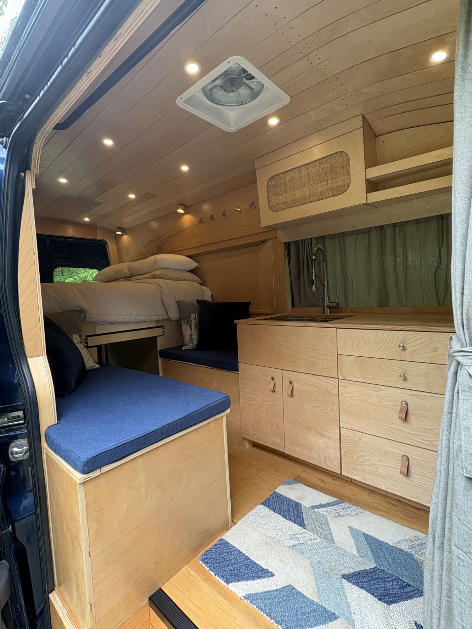 Interior of a small living space or camper van with wooden walls and ceiling, a bed with pillows, a blue cushioned bench, a colorful geometric rug, and a kitchen area with a sink. There are multiple recessed ceiling lights and fabric curtains coverin