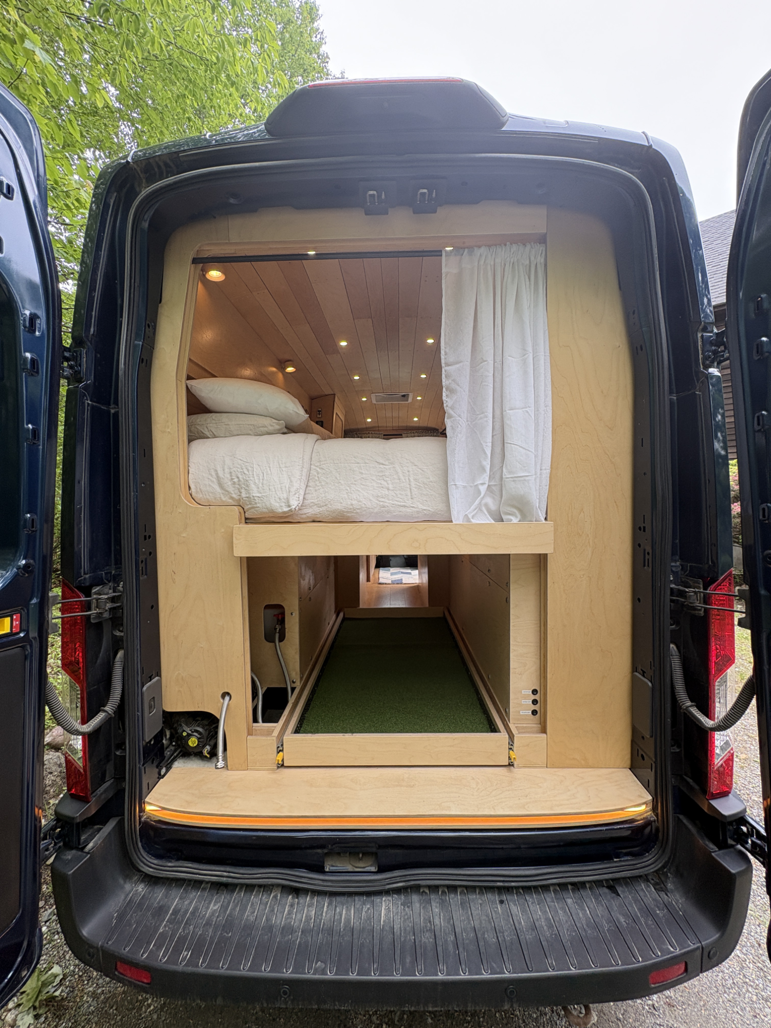 The back of a black van with its rear doors open, revealing a custom-made wooden interior with a bed and storage space.