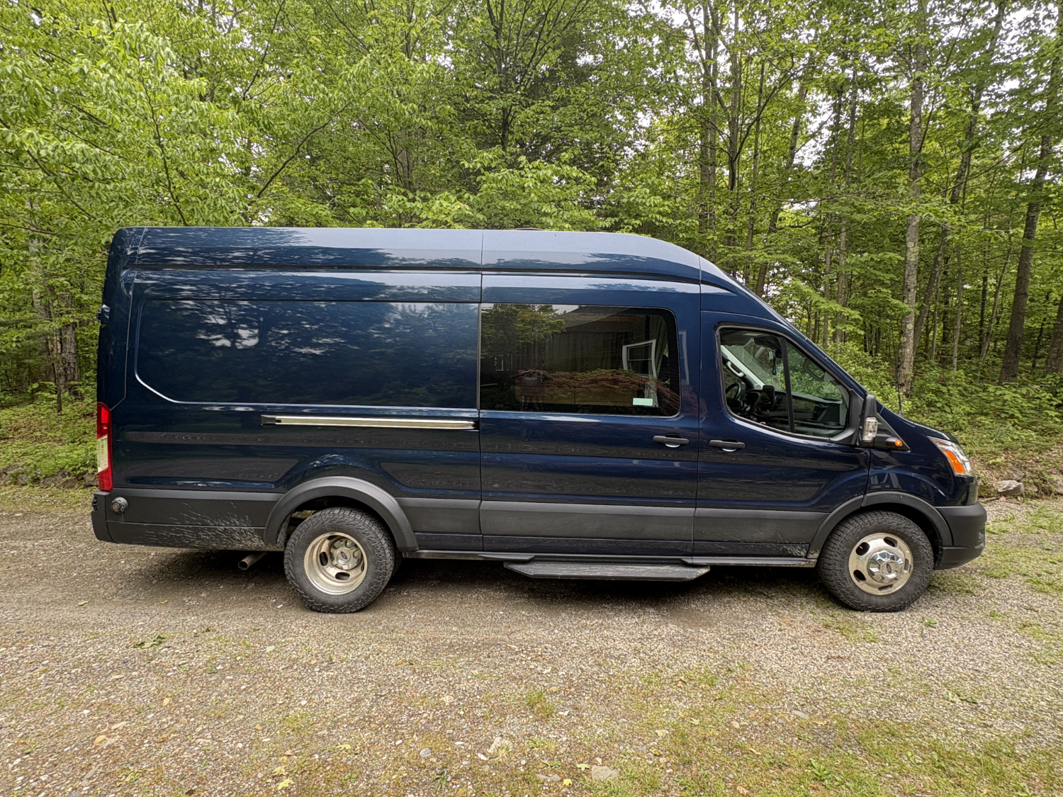 A dark blue cargo van parked on a gravel surface in front of a green forested area.