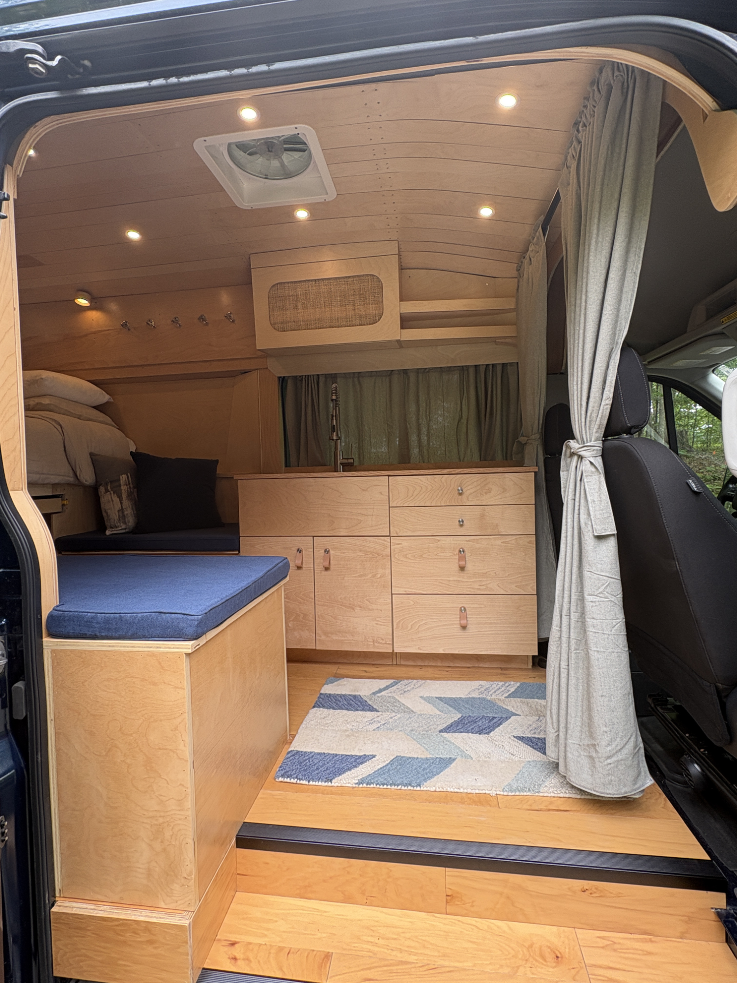 Interior of a small van with wooden interior, including bed, storage cabinets, curtains, and a small rug, with circular ceiling lights.