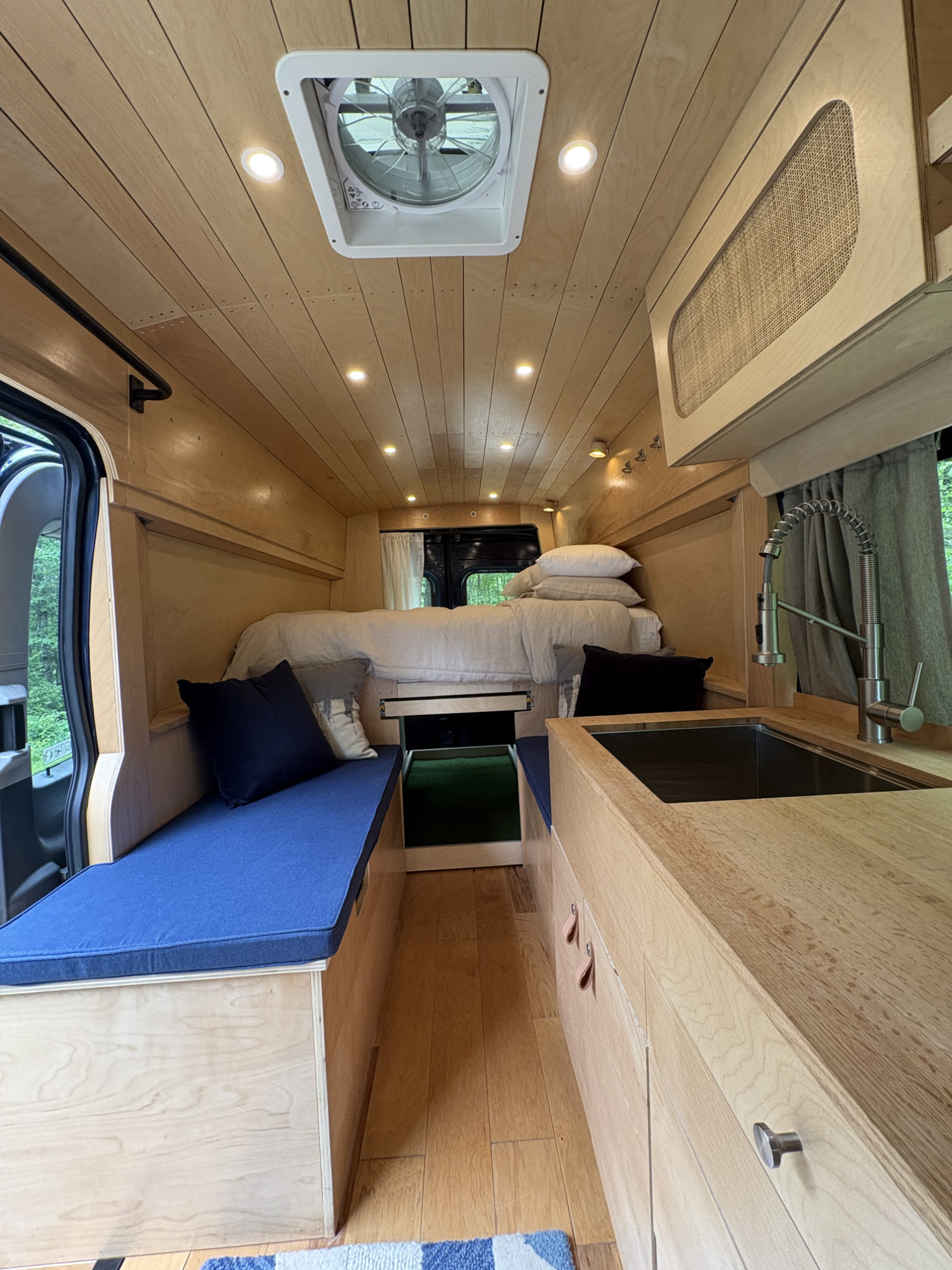 Interior of a tiny home or camper with wooden walls, a bed in the back with pillows, a small kitchen counter with a sink, and a couch with pillows, windows showing greenery outside, and a ceiling vent.