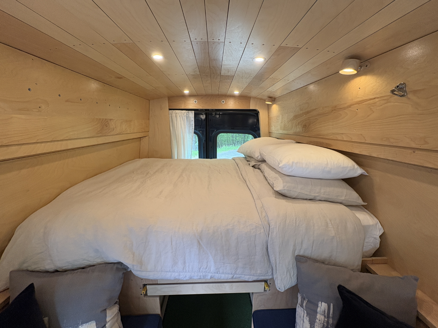 Interior of a wooden camper van with a bed, pillows, and a window with a curtain at the end.