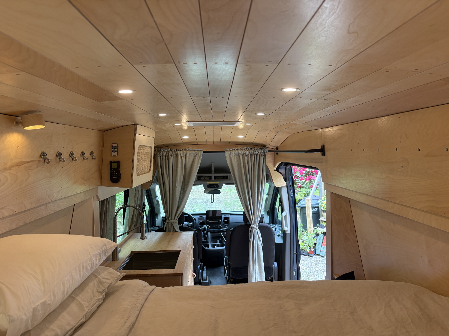 Interior of a converted van with wooden paneling, a bed with pillows, built-in storage, curtains at the driver cab, and view outside showing plants and a garden.