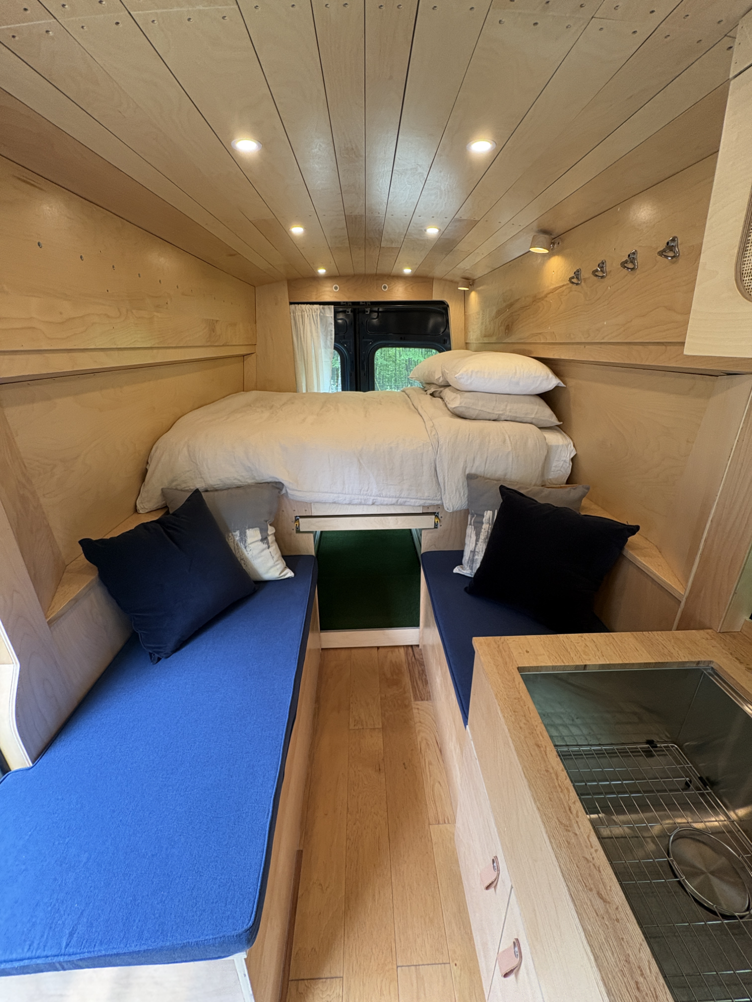 Interior of a small wooden camper with a bed, two blue-cushioned benches, and a small kitchenette area with a sink.