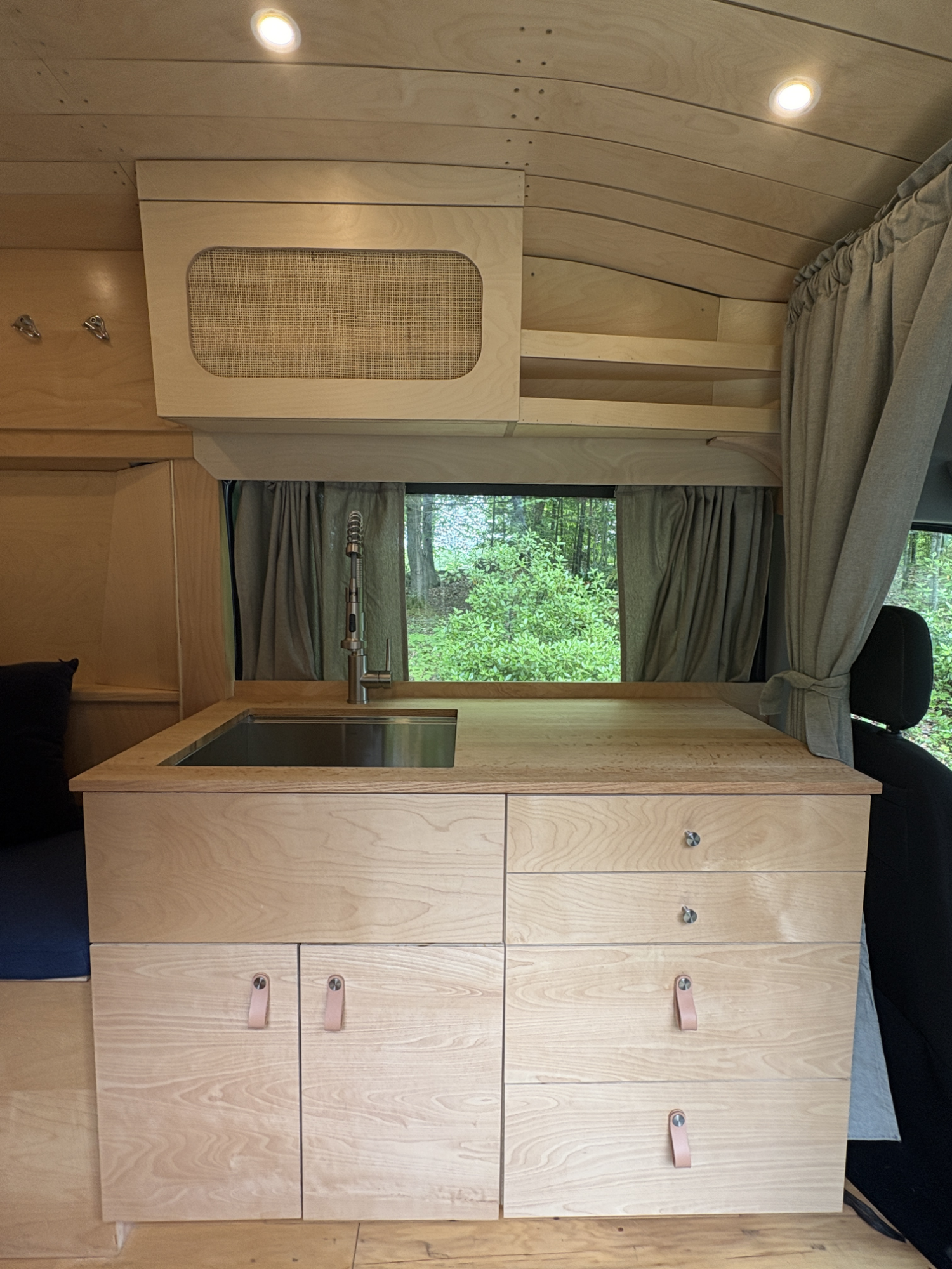 Wooden kitchen unit with a sink, drawers, and shelves, situated in front of a window with curtains, overlooking a green forest.