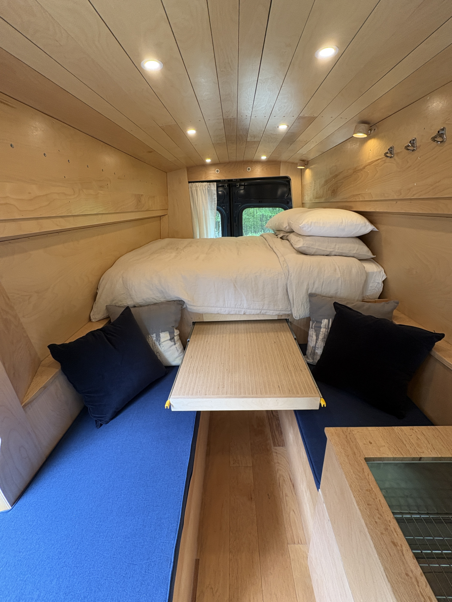 Cozy sleeping area inside a camper van with a bed, pillows, built-in wooden storage, and a small table, illuminated by recessed ceiling lights.