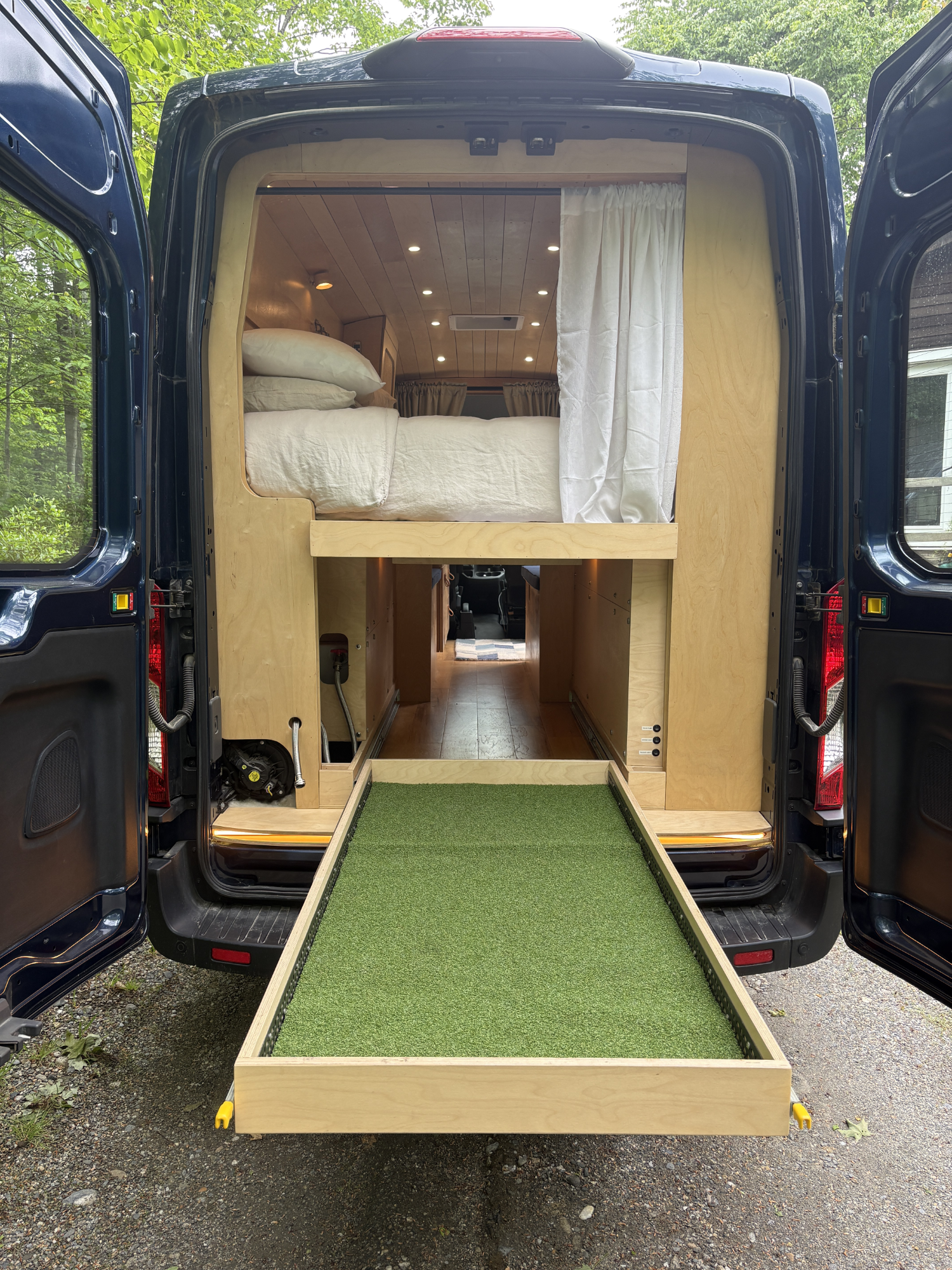 The image shows the back of a van with its doors open, revealing a custom tiny home interior with a bed, curtains, and wooden paneling. There is a green artificial grass ramp leading into the van on the gravel ground outside.