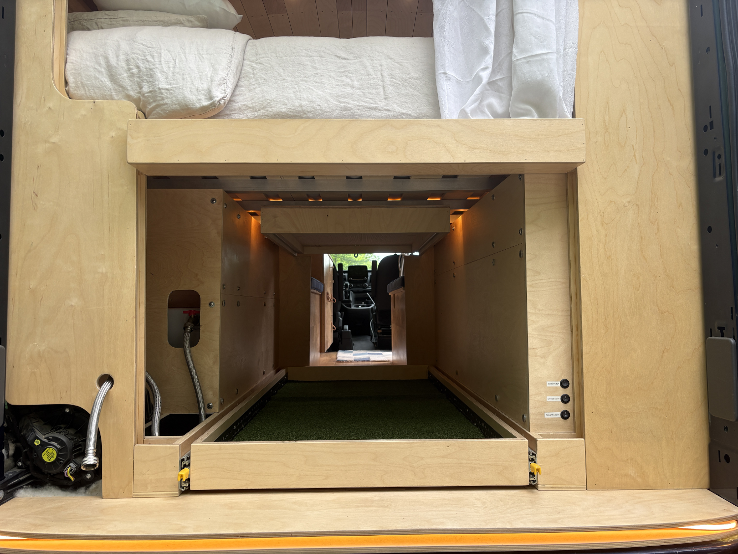 View of the interior of a vehicle with a wooden structure, possibly a custom-built van conversion, showing a bed with a pillow and bedding, plumbing pipes and metal components, and the driver's cabin visible through an opening in the wooden frame.