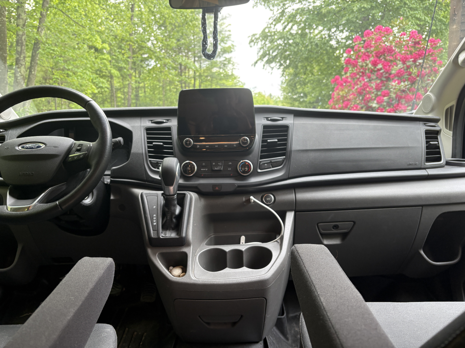 Inside of a vehicle with a black steering wheel, a central dashboard with a screen, gear shifter, and cup holders, with greenery and pink flowers visible outside through the windshield.